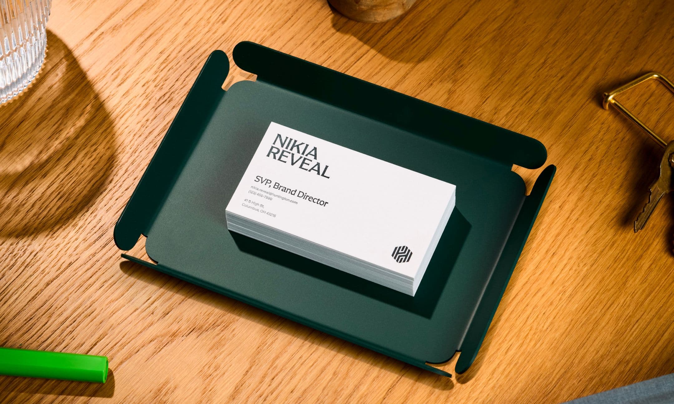 A stack of business cards labeled Nikia Reveal, SVP Brand Director sits on a dark green tray on a wooden desk with office supplies nearby.