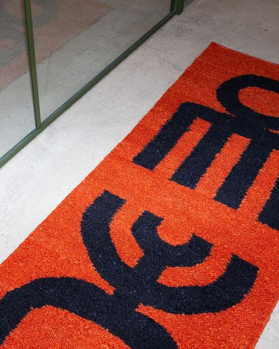 A rectangular doormat with a bright orange background and bold black abstract geometric patterns, placed on a light floor next to a glass wall with a green frame.