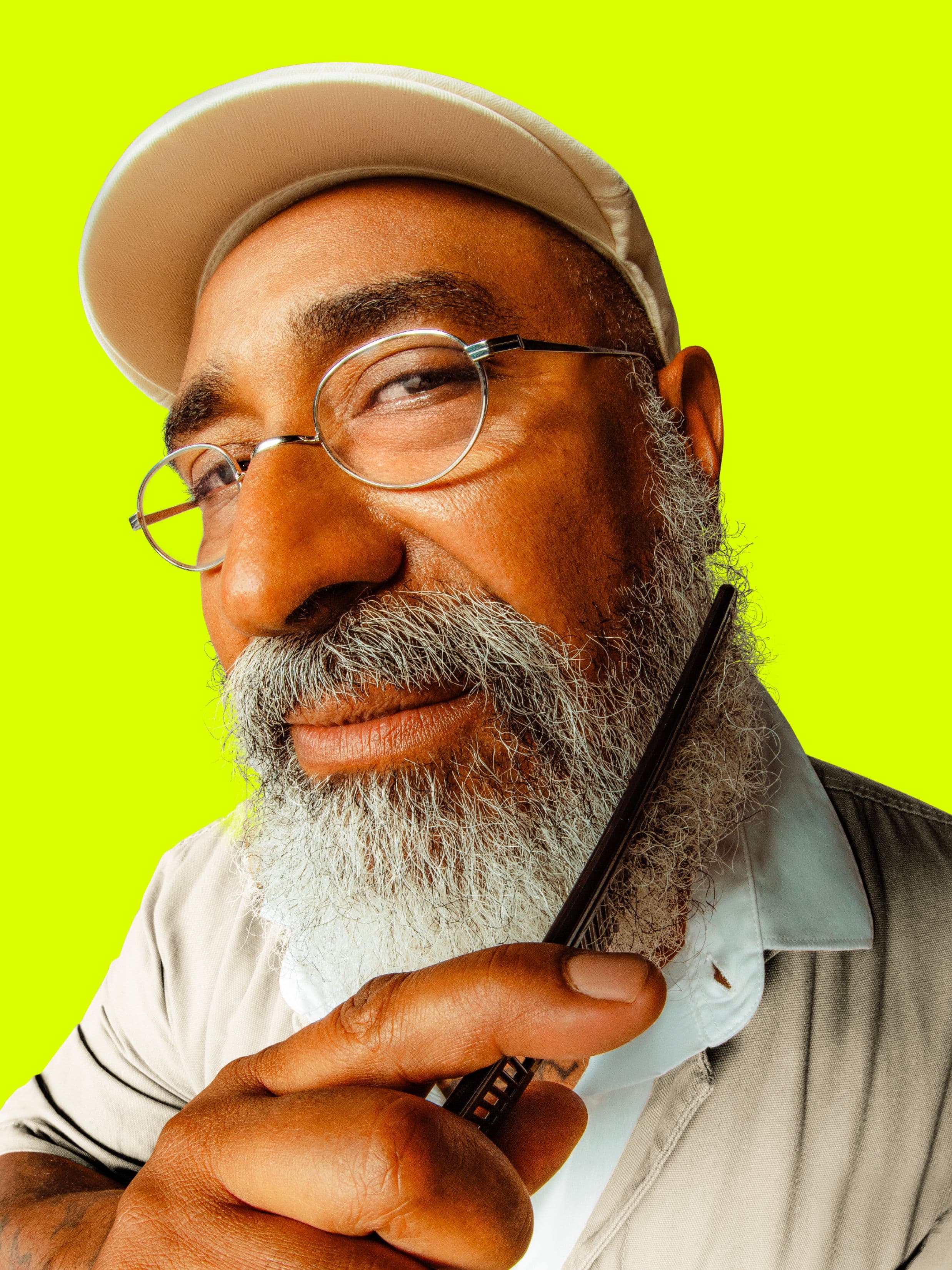 A bearded man wearing glasses and a beige cap smiles slightly while combing his gray beard against a vibrant yellow background.