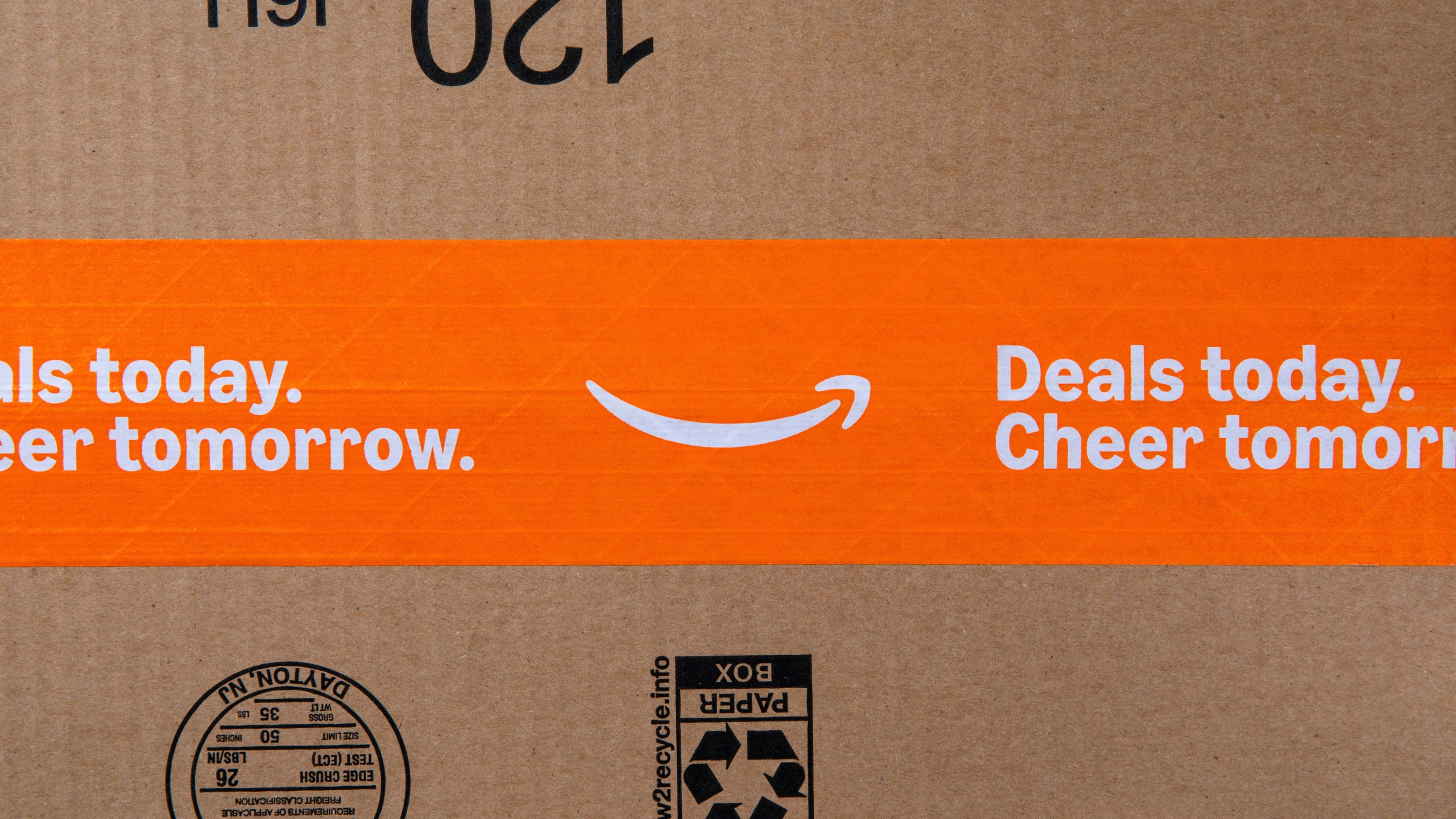A cardboard Amazon box with an orange tape featuring the Amazon smile logo and the text Deals today. Cheer tomorrow. printed in white.