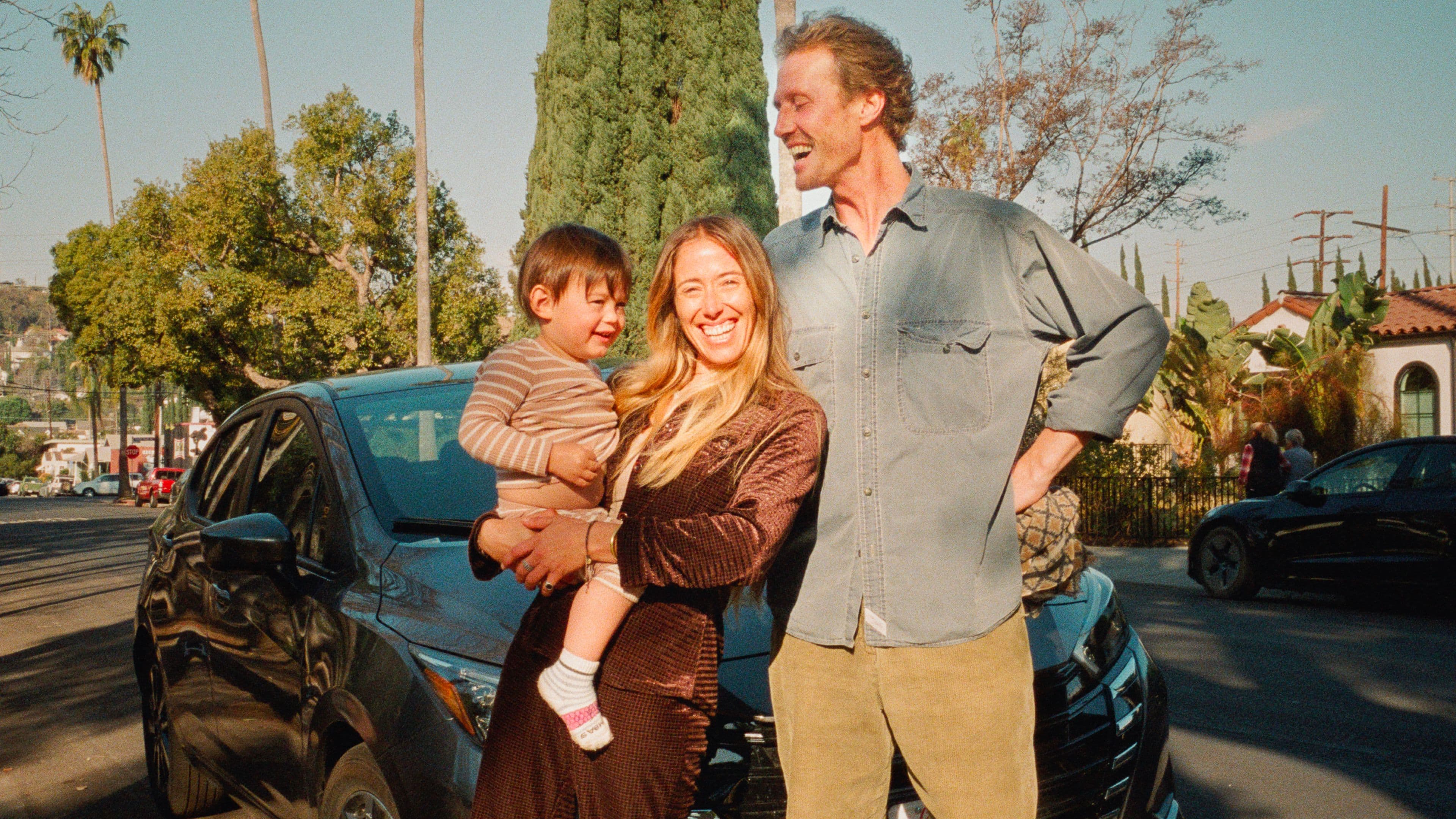 A smiling woman holds a young child, standing next to a man outdoors in front of a black car, with trees and houses in the background on a sunny day.