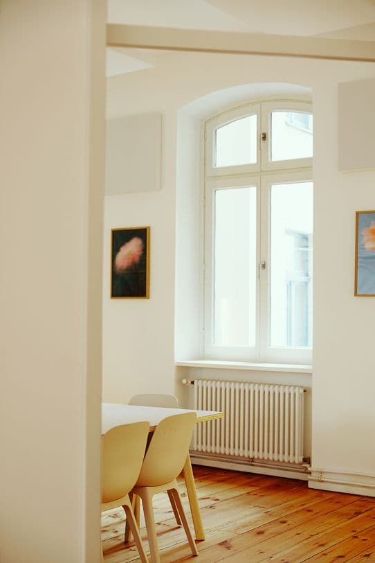 A bright, minimal dining area with a wooden floor, white walls, a large arched window, four light-colored chairs, a white table, and two framed floral pictures on the walls.