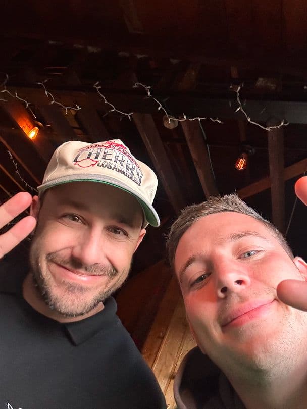 Two smiling men pose for a selfie indoors, both making peace signs.
