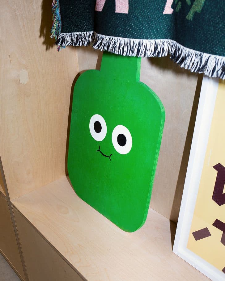 A green, paddle-shaped object with cartoon eyes and a small smile leans against a wooden shelf, partly covered by a fringed, dark-colored fabric.
