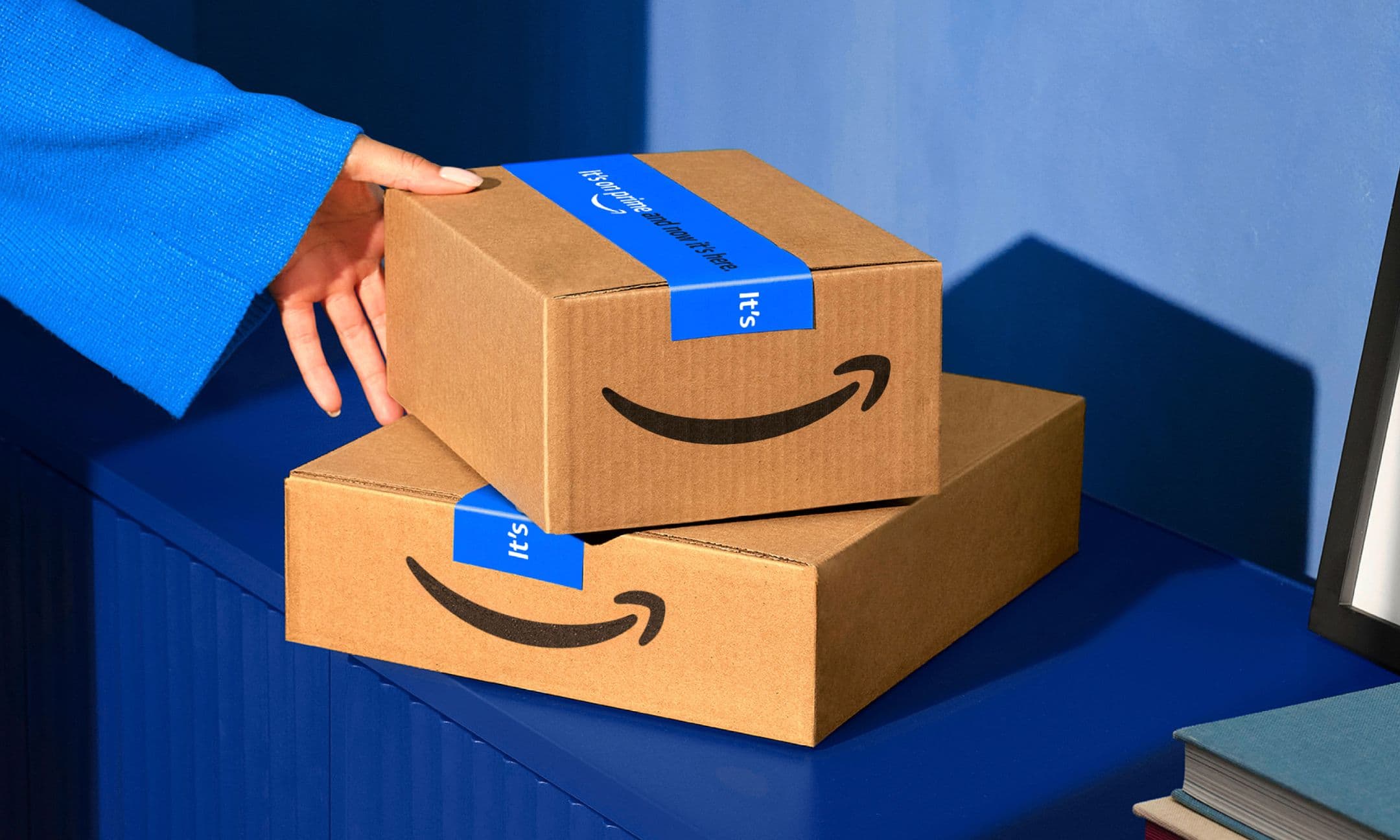 A hand reaches for one of two stacked Amazon delivery boxes, sealed with blue tape, on a blue surface against a blue wall.