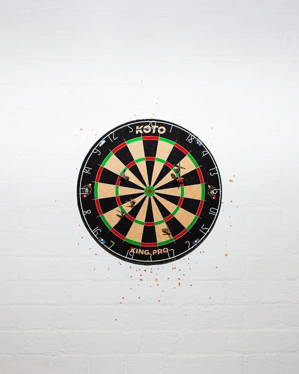 A black, red, and green dartboard is mounted on a white wall, with several darts stuck in it and small marks around the board where darts have hit the wall.