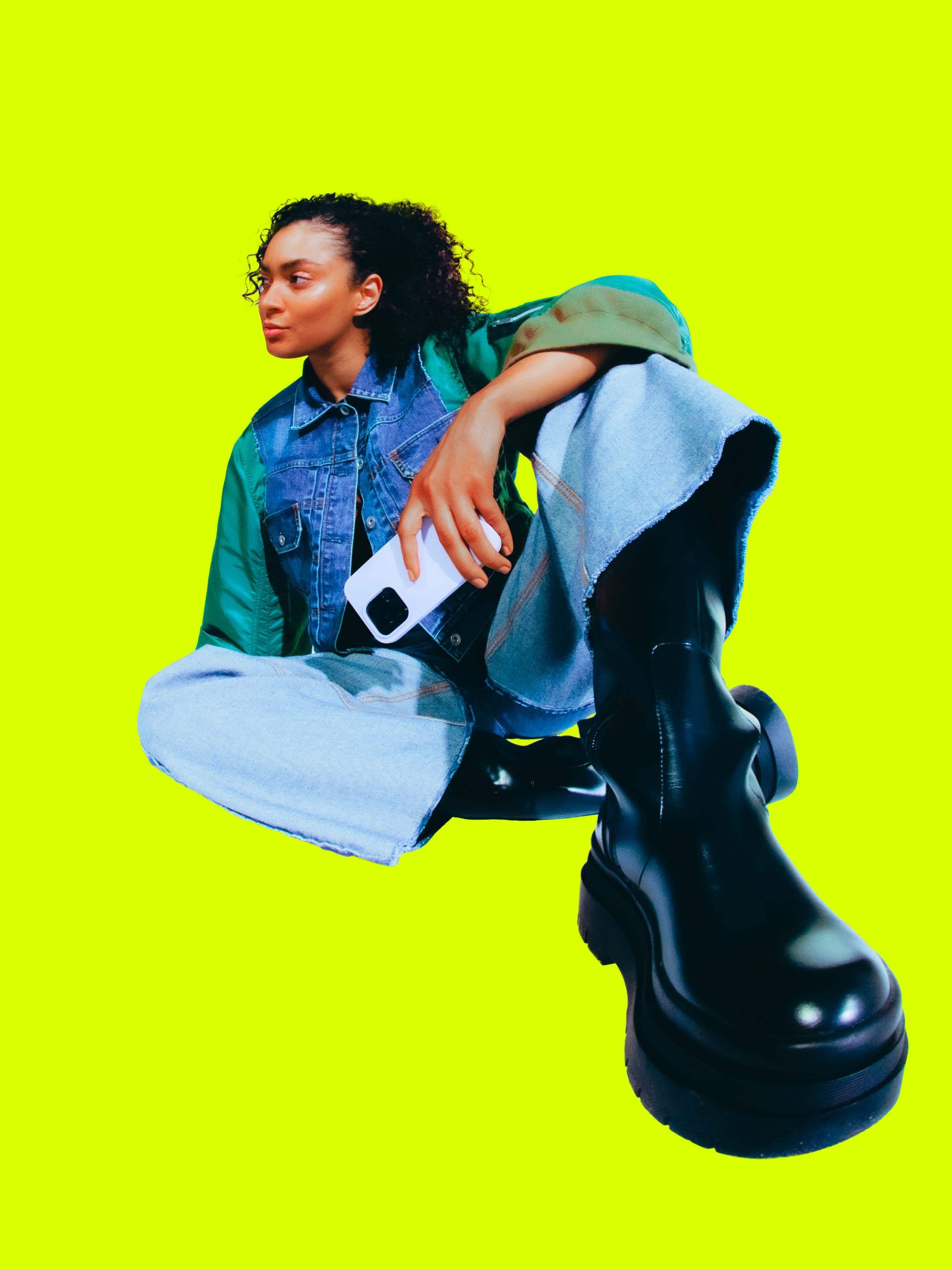 A stylish person in denim and a green jacket sits against a bright yellow background, holding a white camera. The photo is taken from a low angle, emphasizing their large black boots.