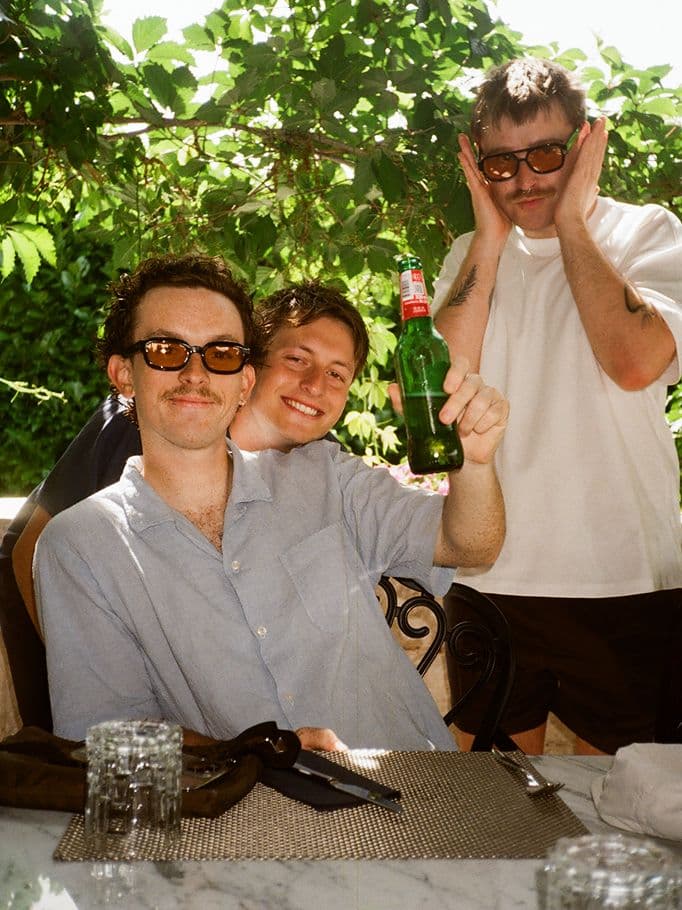 Three men pose at an outdoor table; one in a blue shirt holds up a green beer bottle and smiles, another leans in and grins, while the third, in sunglasses, stands behind them with hands on his face, all enjoying a sunny day.