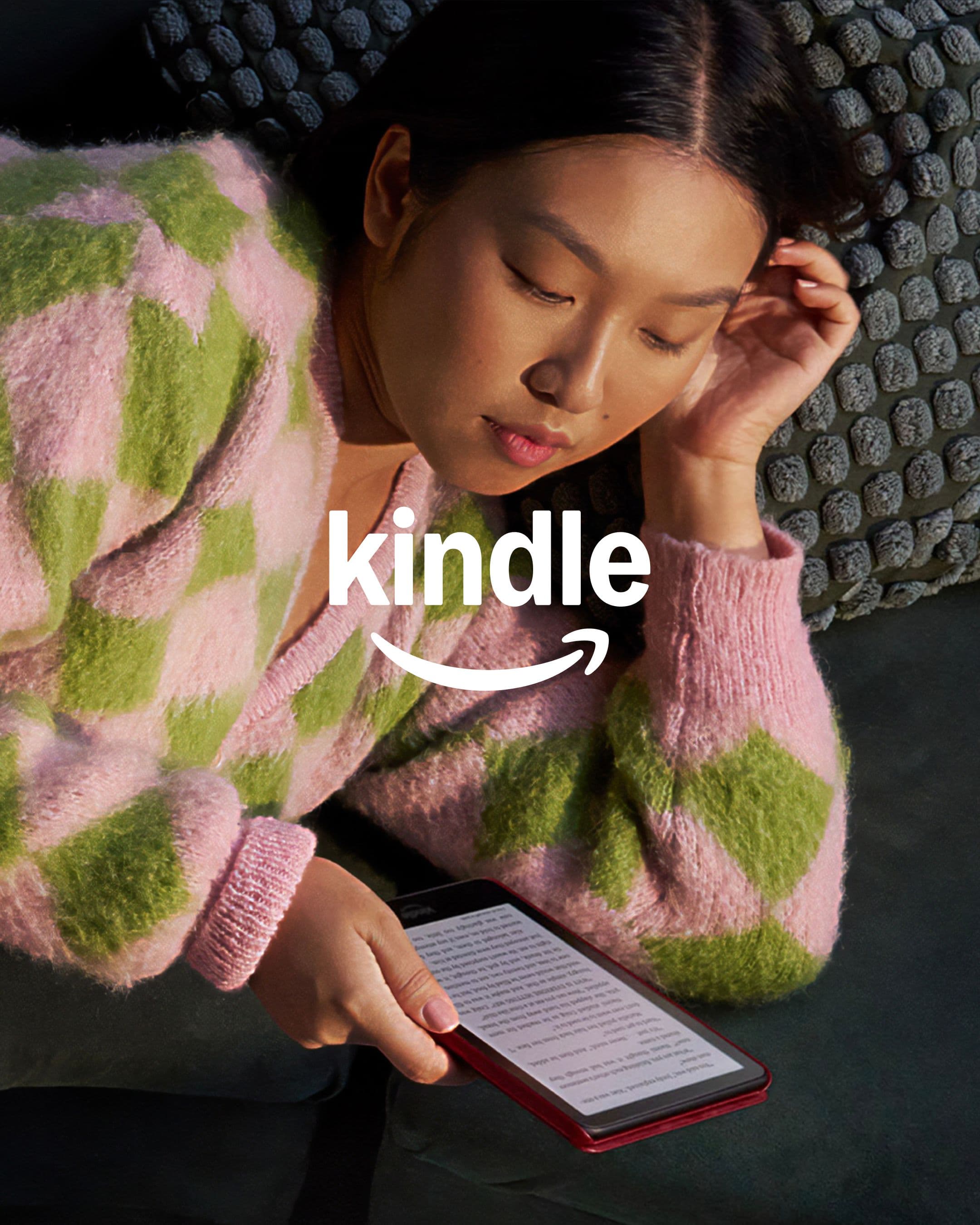 A person wearing a pink and green checkered sweater is lying on a couch, reading an e-reader. The word kindle with the Amazon smile logo below.