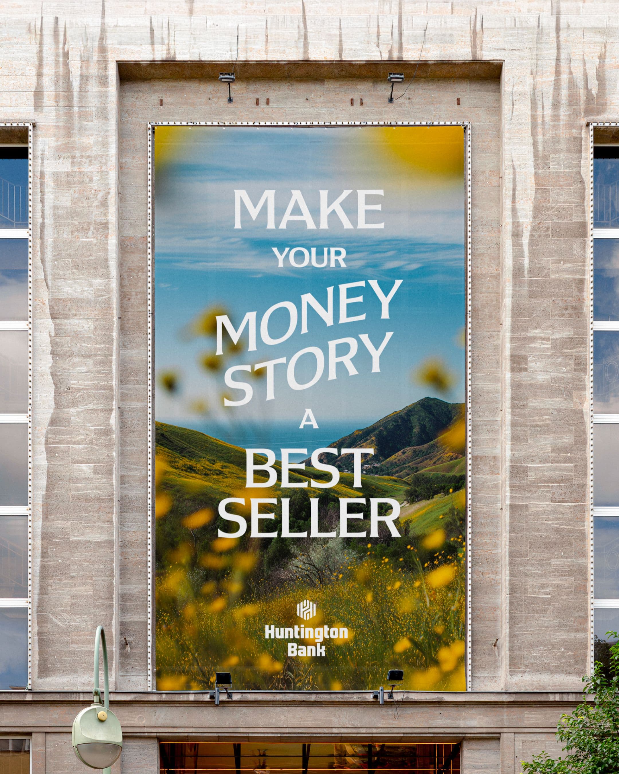 A large outdoor banner on a stone building shows a scenic landscape with green hills and yellow flowers, displaying the text: MAKE YOUR MONEY STORY A BEST SELLER. Huntington Bank with the bank’s logo.