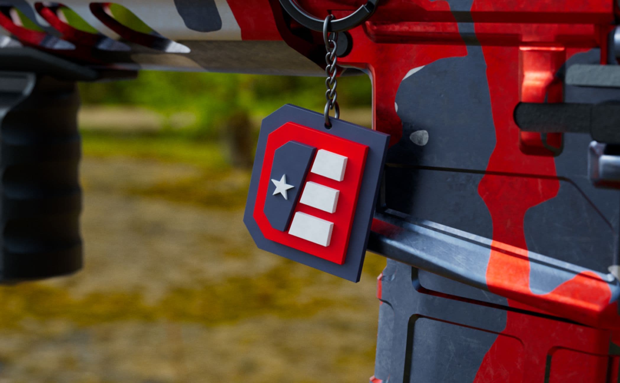 A close-up of a red, black, and gray camouflage firearm with a keychain attached, displaying a red and black tag featuring a white star and three white bars on a red background. The background is blurred greenery.