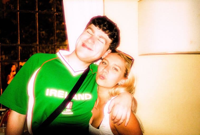A young man in a green Ireland shirt smiles with his eyes closed and arm around a young woman with blonde hair, who is pouting playfully. They stand indoors near a window and a bright lamp.