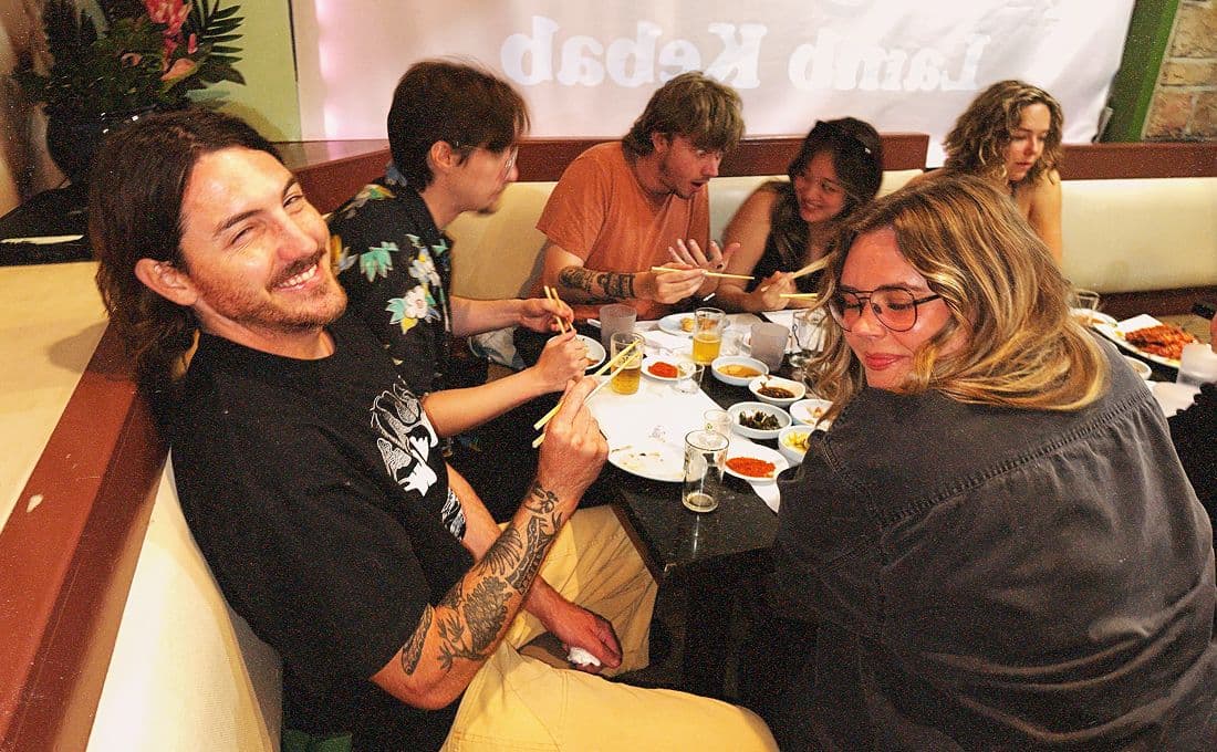 A group of six young adults sits around a restaurant table, smiling and talking while eating. One man in the foreground grins at the camera, holding chopsticks, while others are engaged in conversation and enjoying their meal.