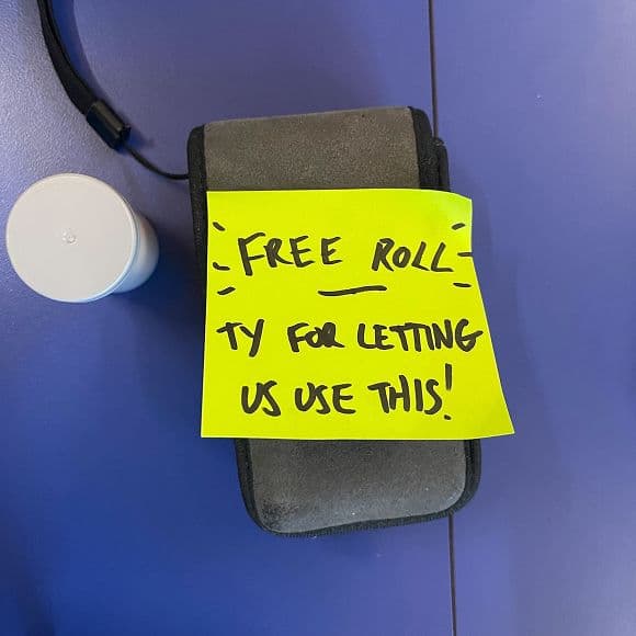 A gray camera bag with a yellow sticky note reading FREE ROLL — TY FOR LETTING US USE THIS! sits on a blue surface next to a white film canister.