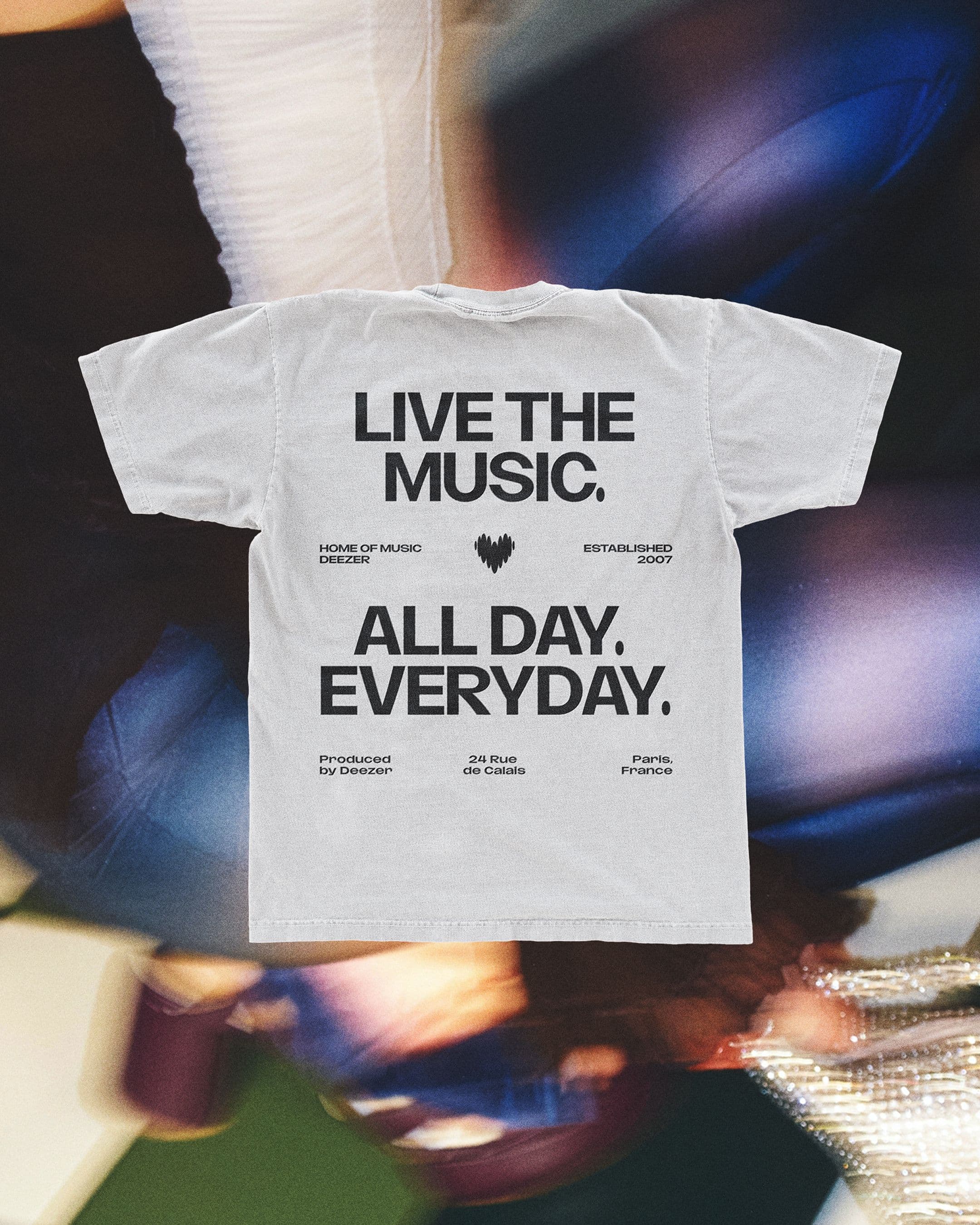 A white t-shirt with bold black text reading, LIVE THE MUSIC. ALL DAY. EVERYDAY. on a shaky image background.