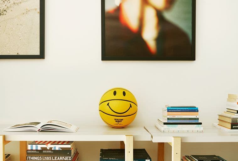 A yellow basketball with a smiley face sits on a shelf between stacks of books; two framed pictures hang on the wall above the shelf.