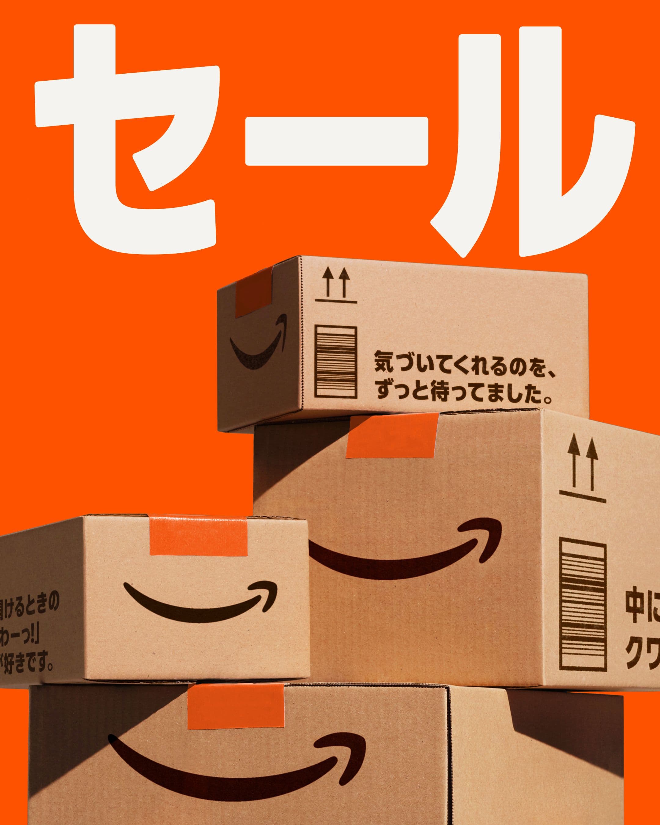 Four Amazon delivery boxes are stacked in front of a bright orange background. Large white Japanese text above reads “セール,” meaning “sale.” The Amazon smile logo is visible on each box.