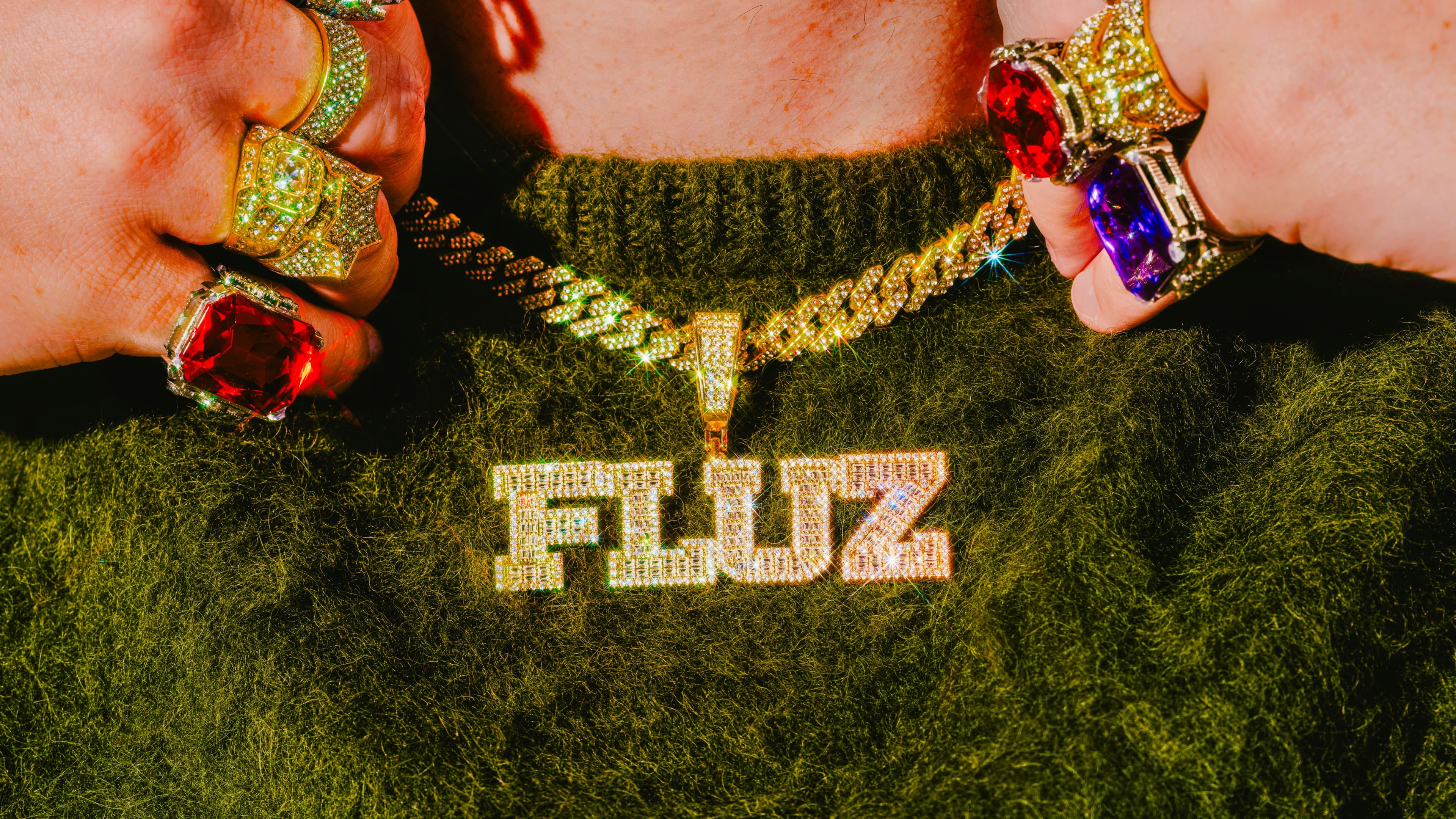 A person wearing a fuzzy green sweater displays a gold chain necklace with a large, sparkling pendant that reads FLUZ. Both hands are adorned with flashy, colorful gemstone rings.