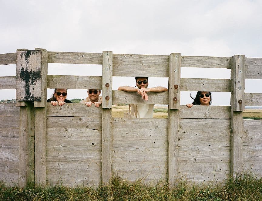 Four people are peeking through rectangular openings in a wooden fence, each at a different height. They are outdoors on a grassy area and wearing sunglasses, with a cloudy sky in the background.