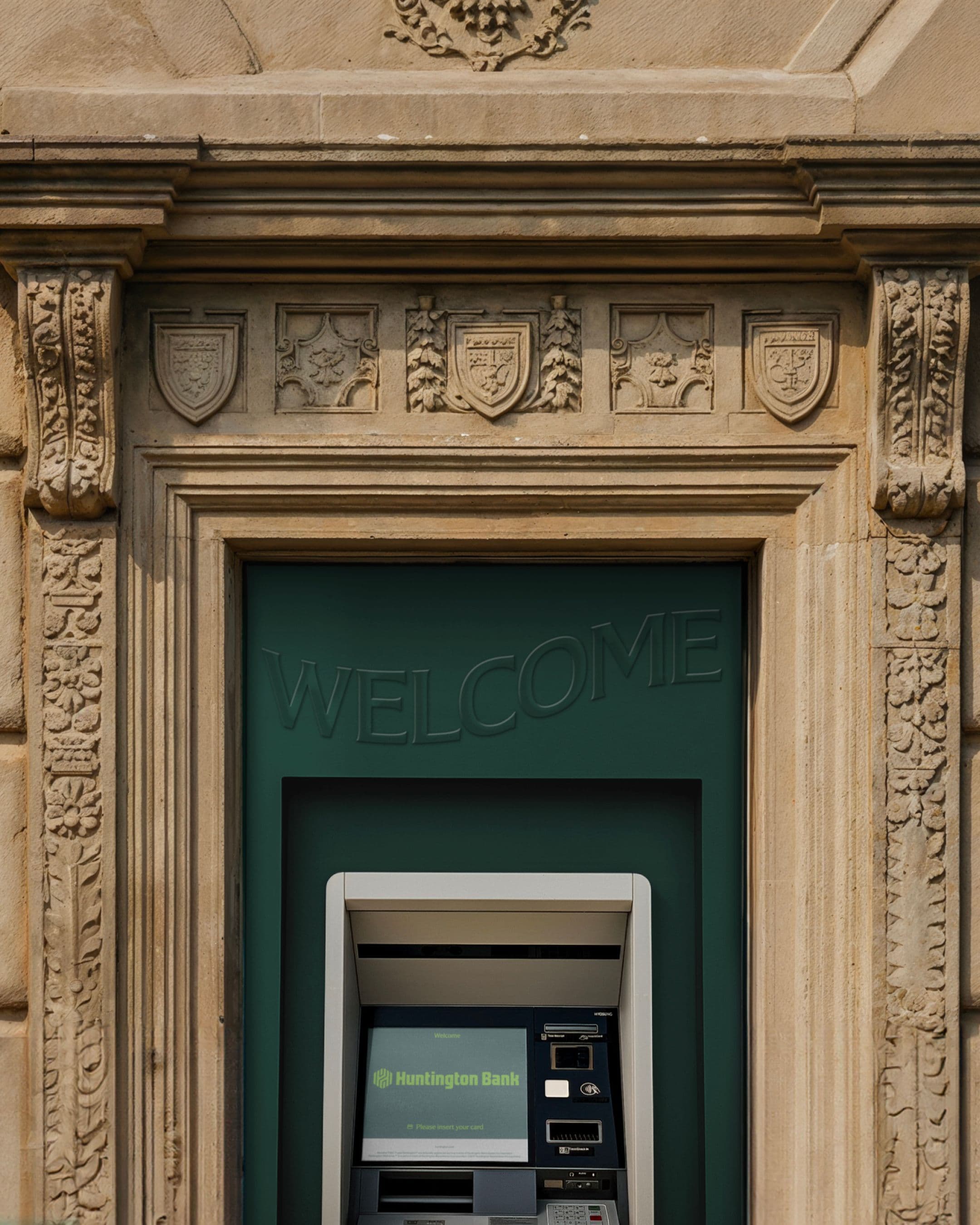 An ATM labeled “Huntington Bank” is set into an ornate stone wall with carved shields and decorative patterns. The word “WELCOME” is displayed above the machine on a green panel.