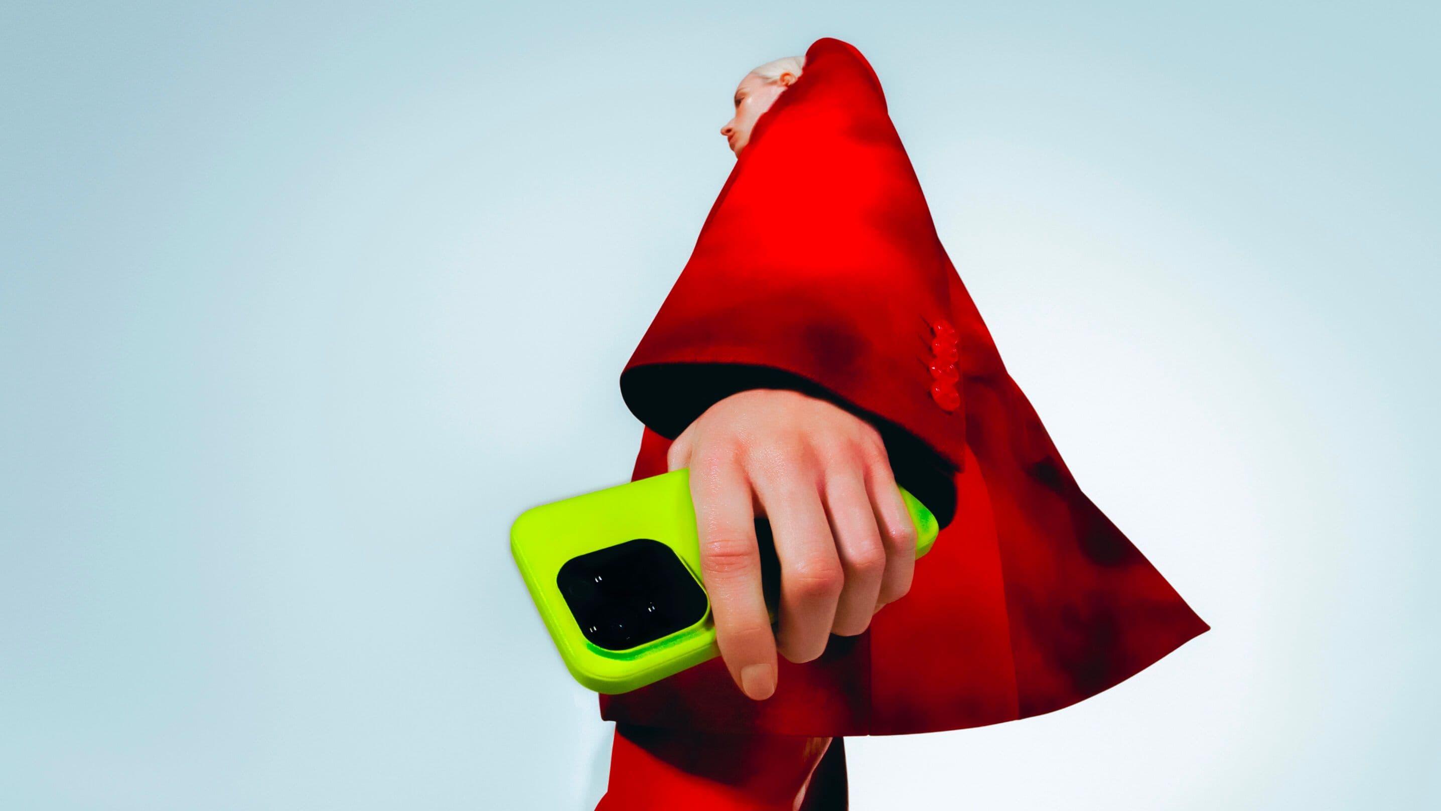 A person in a bold red coat holds a bright green smartphone, seen from a dramatic low angle against a plain, light background.