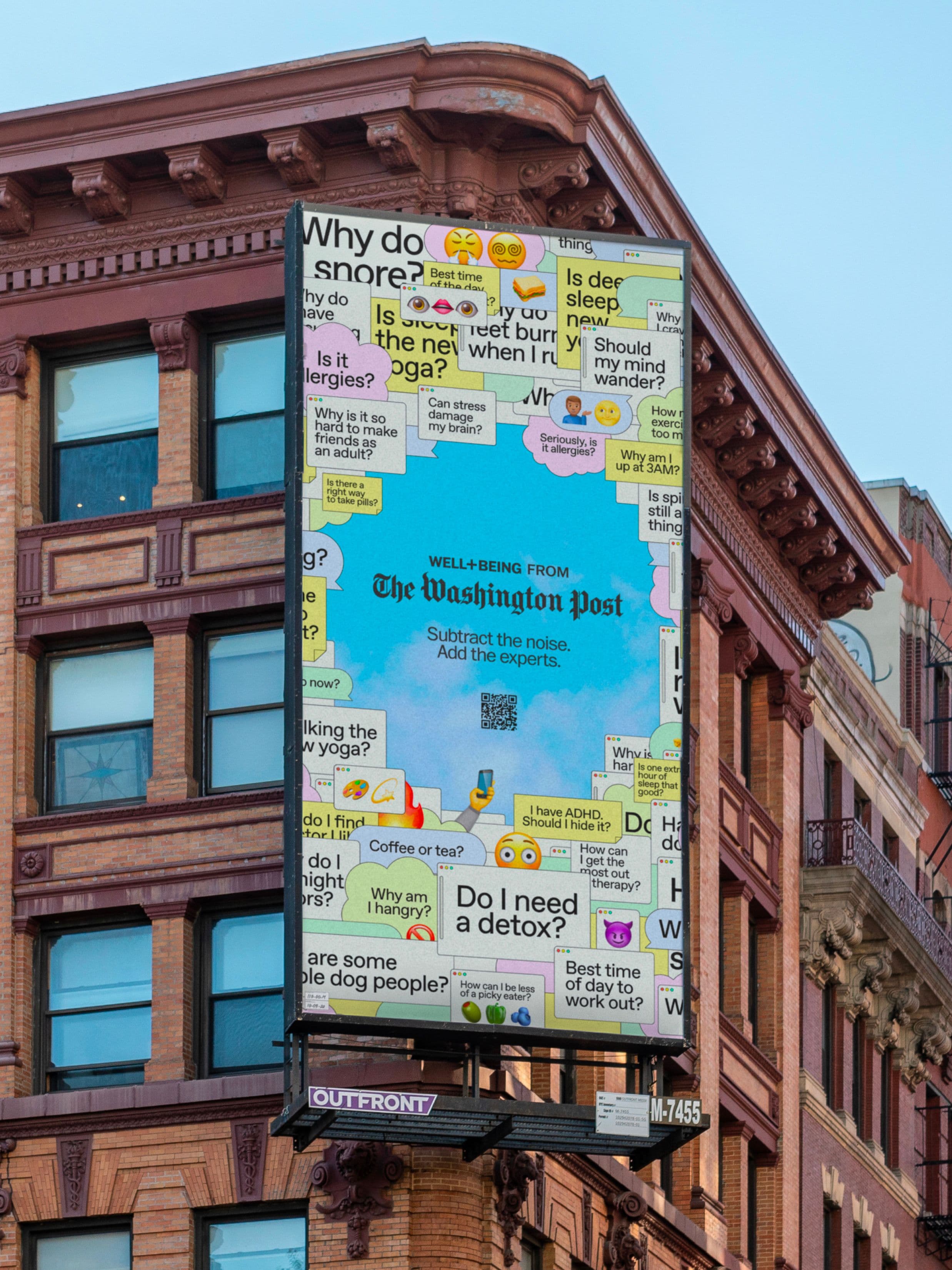 A large billboard for The Washington Post’s well-being section covers the side of a brick building, surrounded by colorful question bubbles about health, wellness, and emojis on a bright blue background.