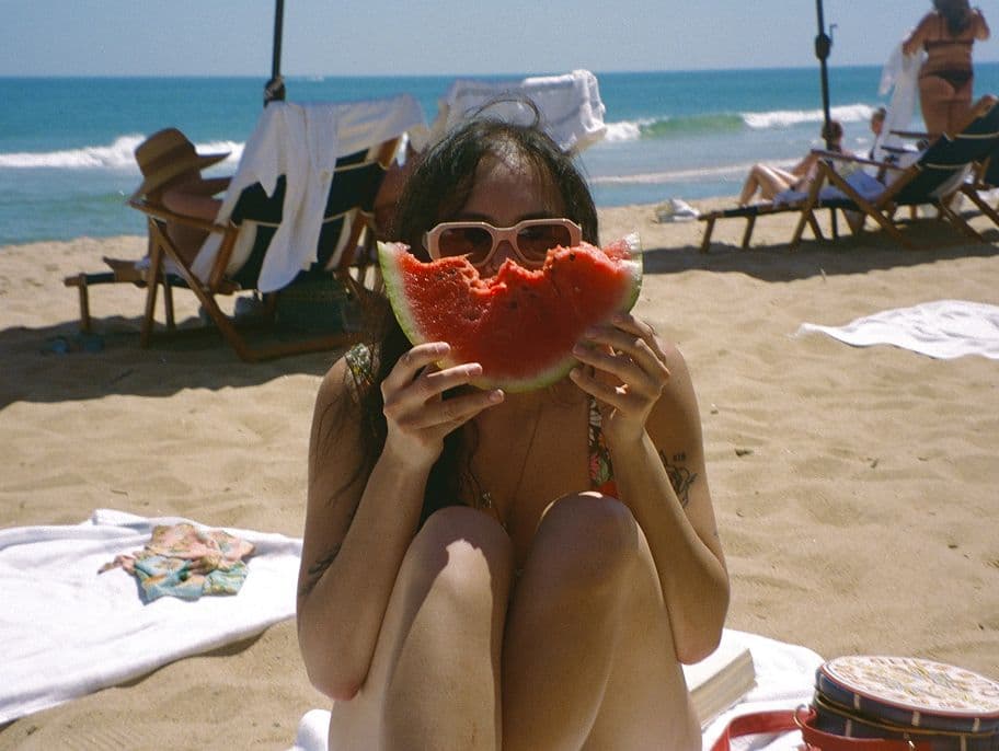 A woman sitting on a sandy beach holds a large slice of watermelon, partly covering her face. She wears sunglasses and is surrounded by beach chairs, towels, and people relaxing near the ocean.