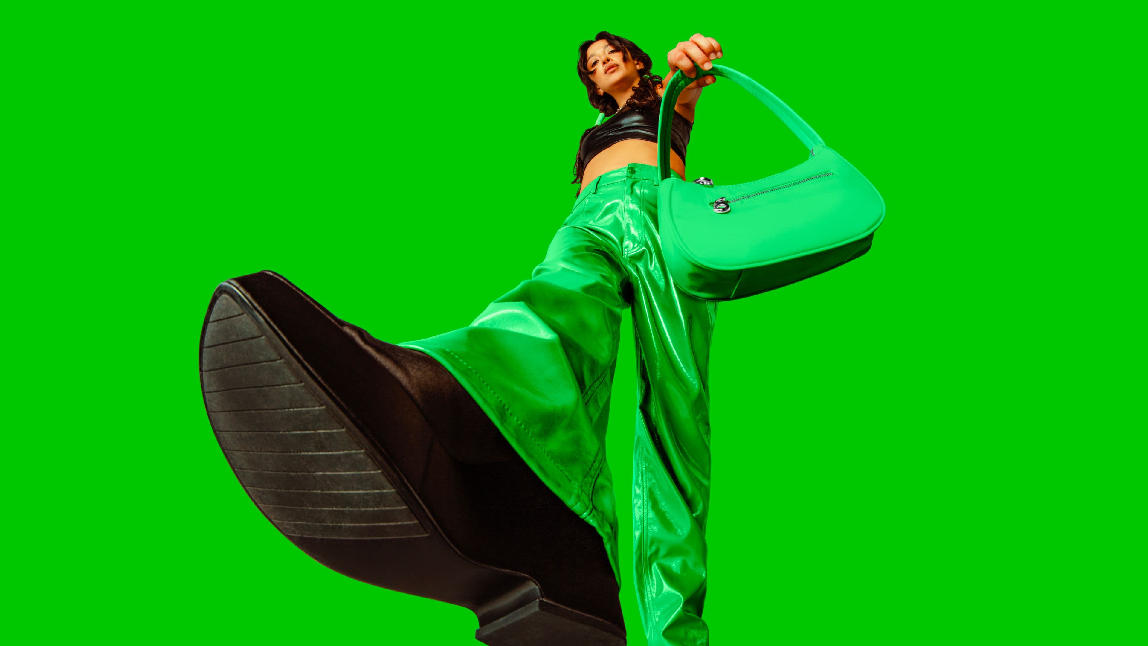 A woman poses confidently against a bright green background, wearing shiny green pants and black platform shoes, holding a matching green handbag toward the camera from a low angle.