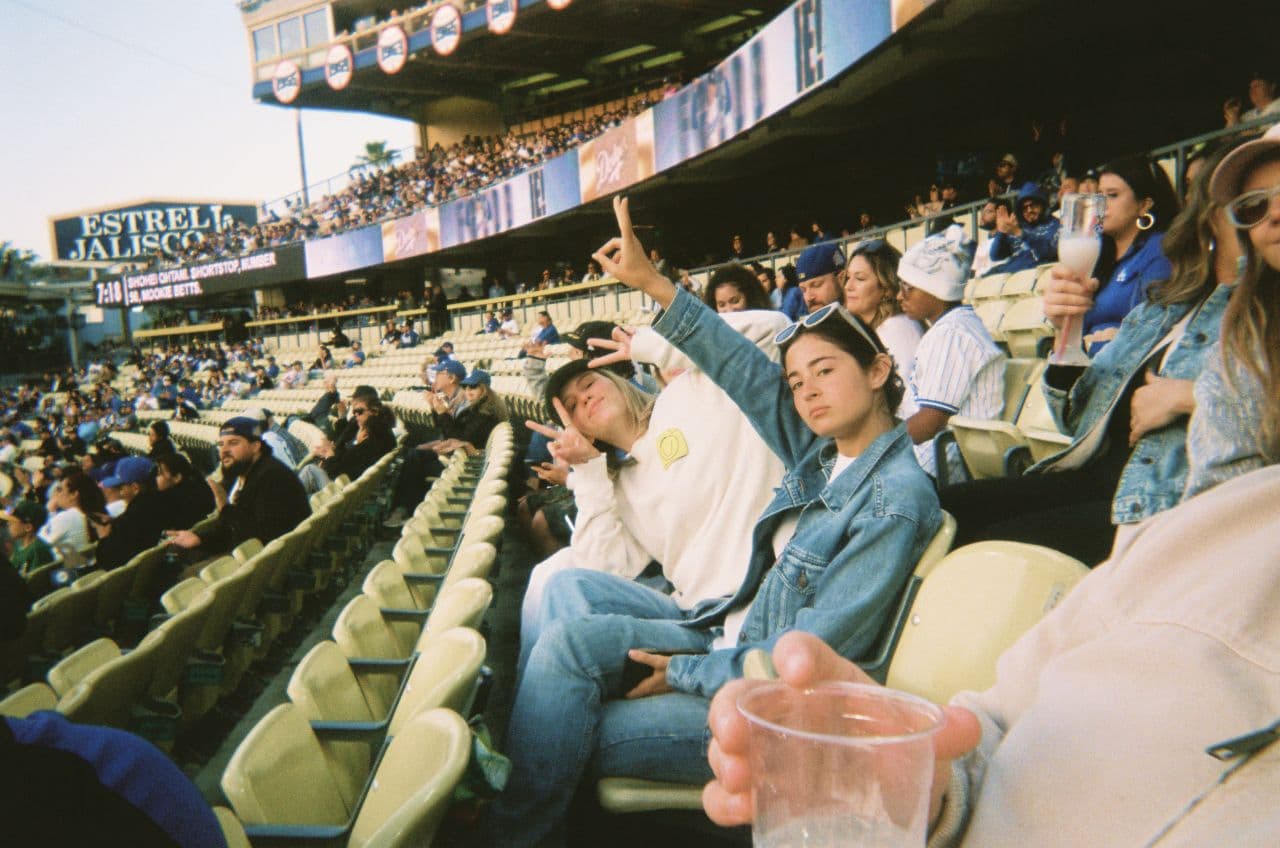 Two people sit in stadium seats, posing and making peace signs at the camera. Other spectators are around them, some wearing baseball caps and holding drinks. The stands and a large advertisement board are visible in the background.