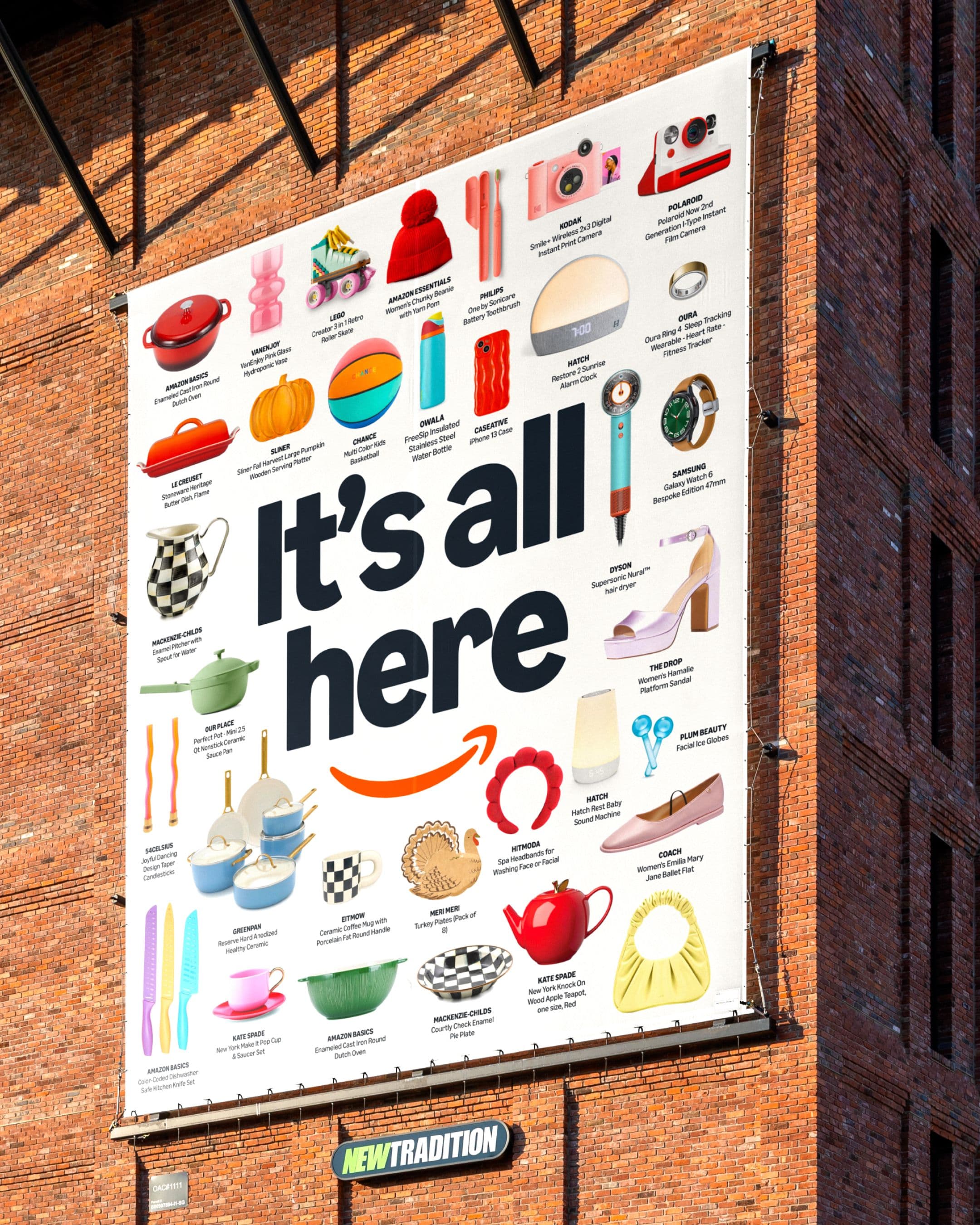 Outdoor billboard on a brick building reading ‘It’s all here,’ surrounded by product imagery and the Amazon smile arrow logo.
