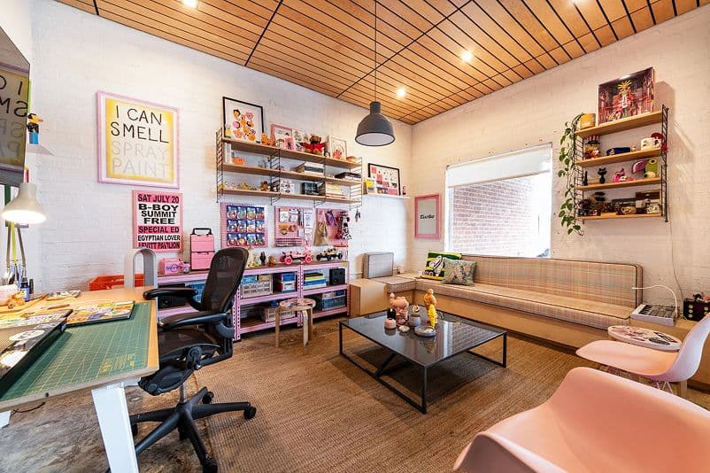Colorful, eclectic office with art supplies, posters, and toys on shelves. There’s a plaid sofa, pink chairs, a central coffee table, and a desk with a green cutting mat. The room has wood ceilings and a cozy, creative vibe.