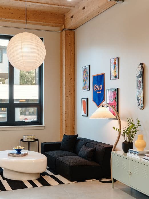 Modern living room with a black sofa, white oval coffee table, striped rug, and wall art above the couch. A floor lamp and a side cabinet with plants and decor are also visible under a large paper pendant light.