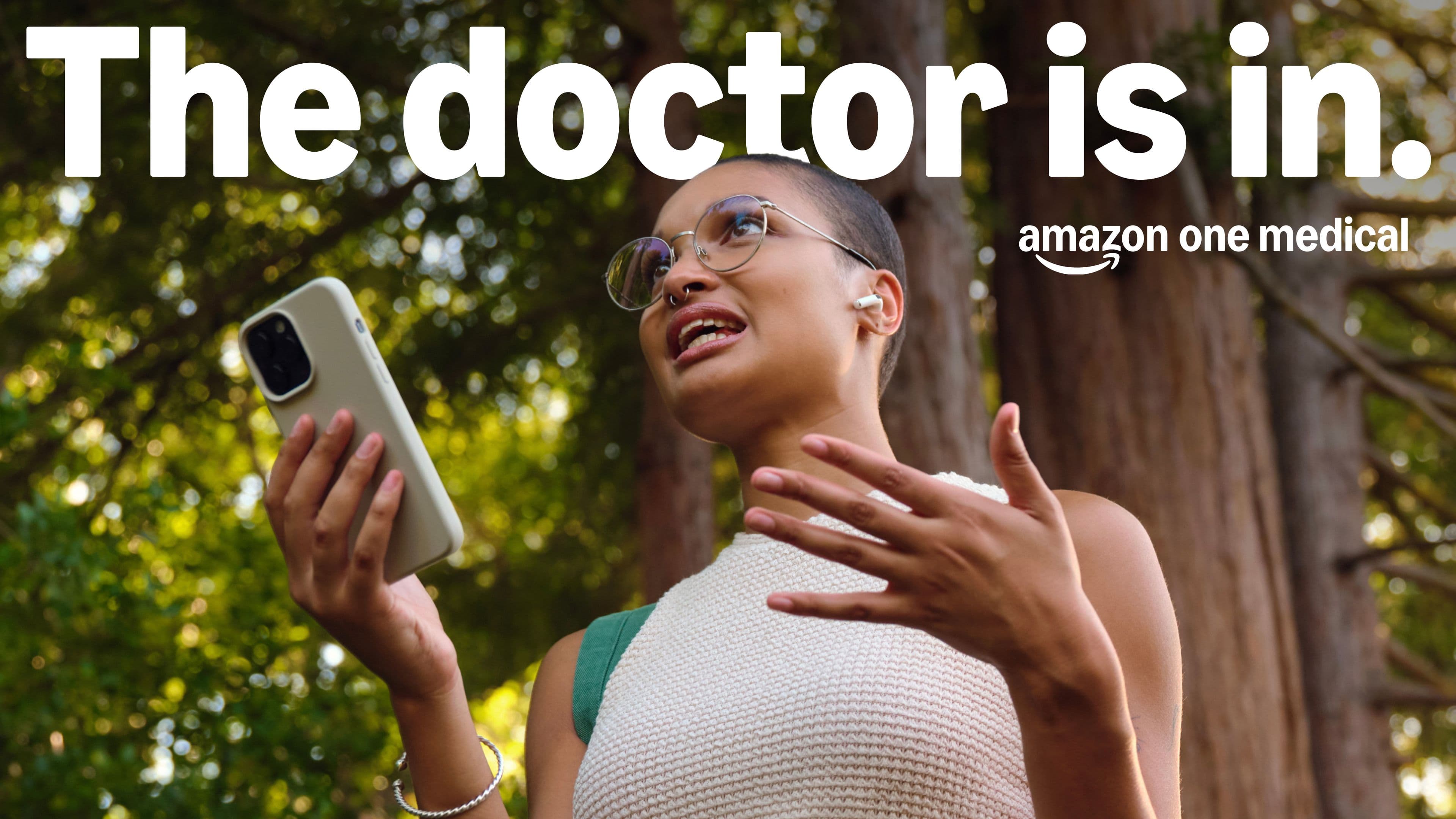 A person with glasses holds a smartphone and gestures while speaking outdoors. Large text reads “The doctor is in.” with the Amazon One Medical logo below.