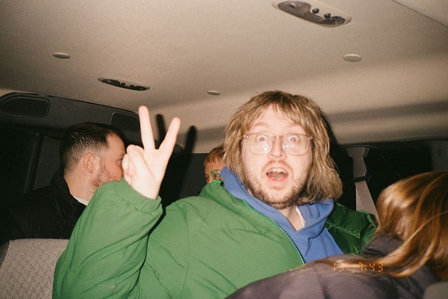 Koto employee in a vehicle wearing a green jacket and blue hoodie, making a peace sign with a surprised expression.