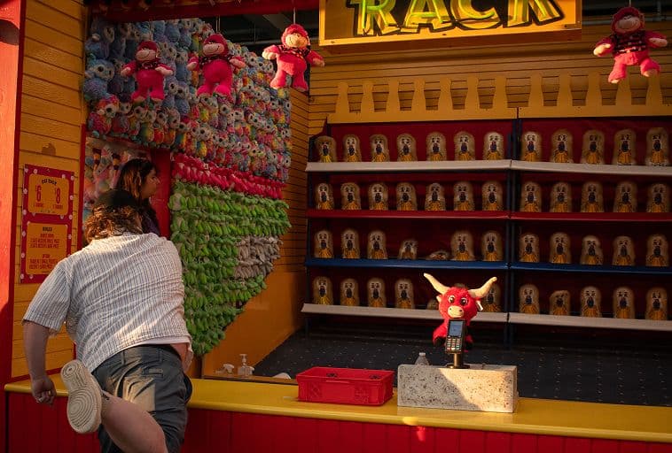 A person stretches their leg at a carnival game booth with plush toys as prizes on the shelves and walls. Bright red and yellow colors decorate the booth, and a red bull plush is displayed on the counter.