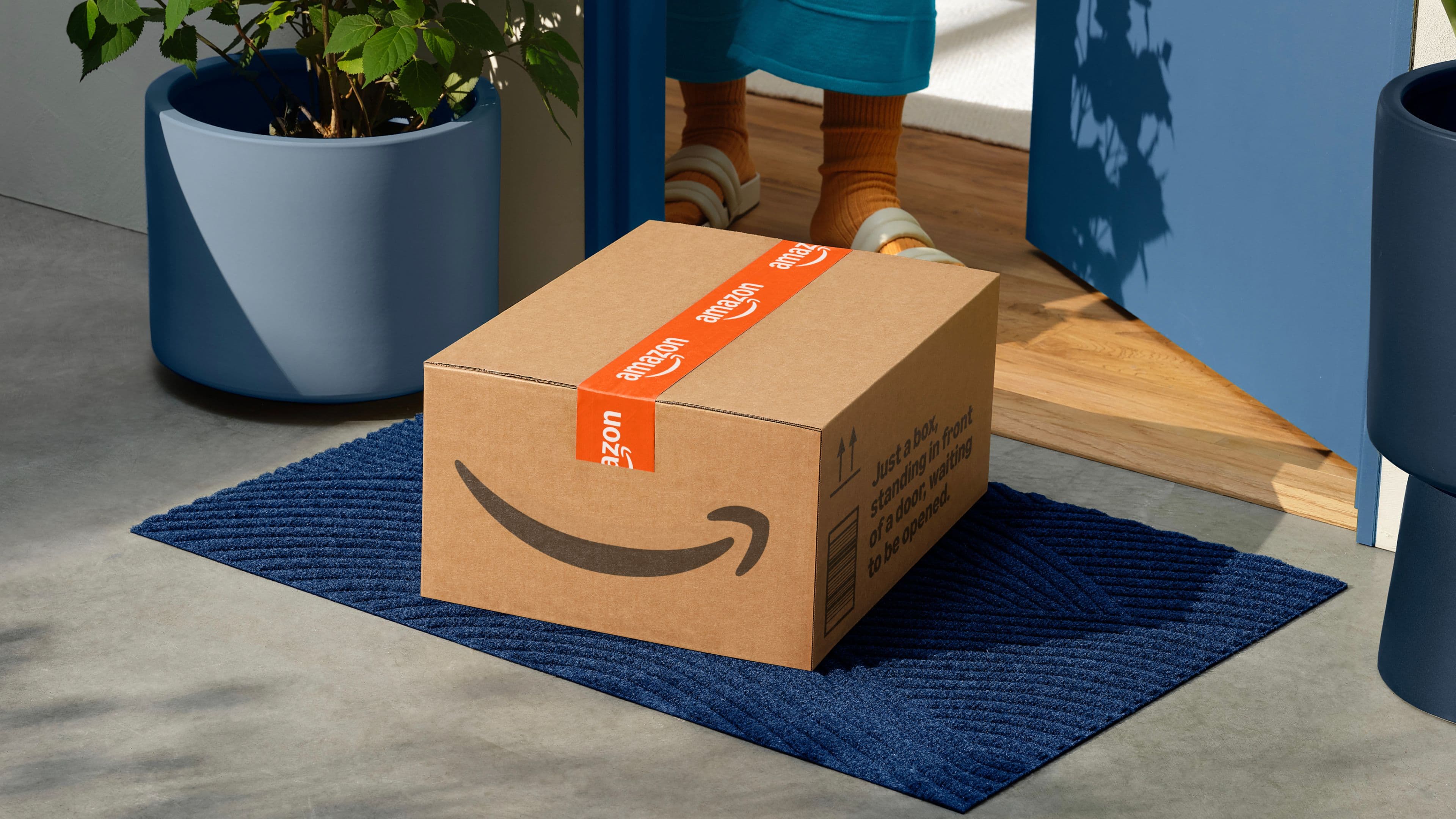 A cardboard Amazon package sits on a blue doormat in front of an open door.