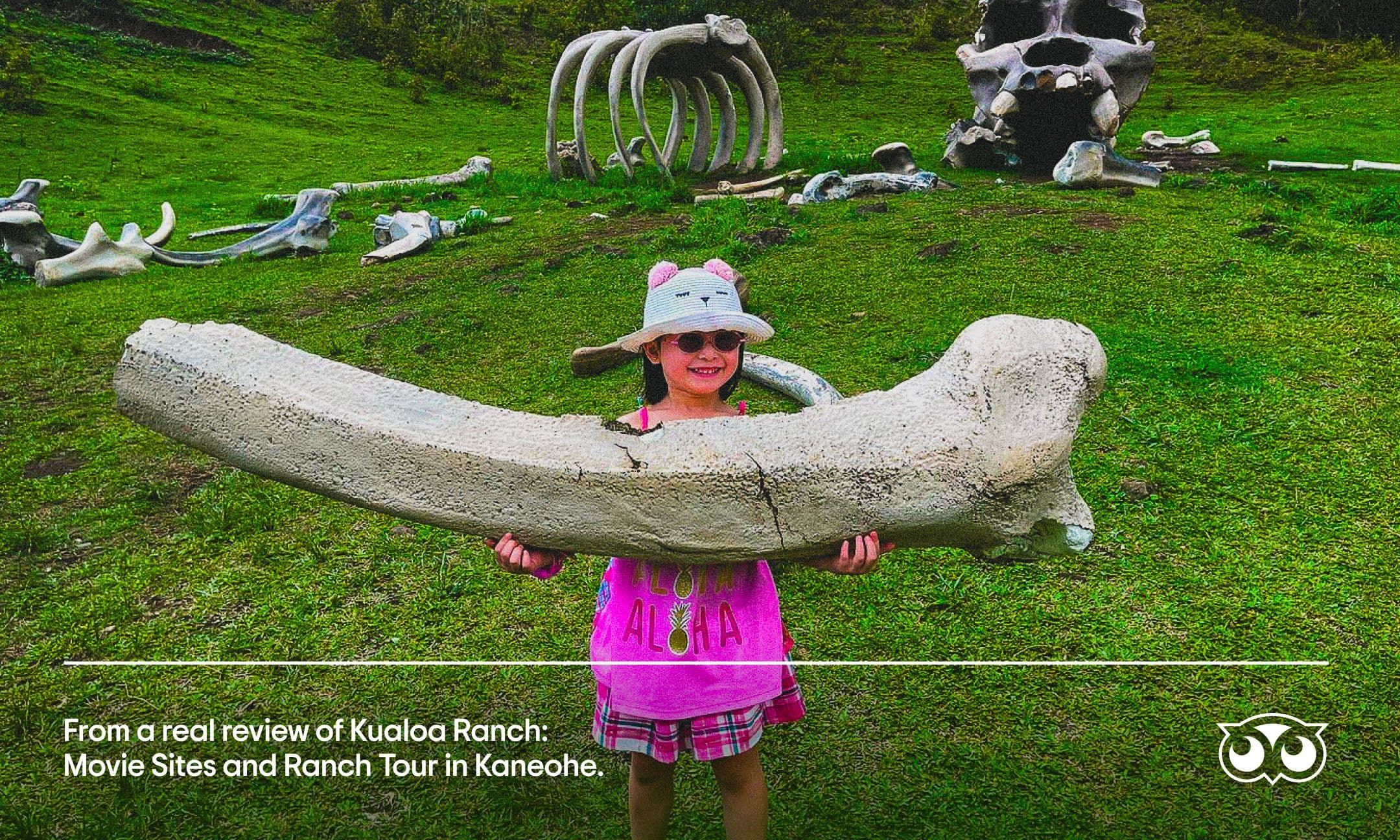 Tripadvisor ‘Seeing the Real View’ campaign: A child in a pink dress, sunglasses, and a cat-eared hat smiles while holding a large fake bone on a grassy field with giant bones and fossil props at Kualoa Ranch, Kaneohe.