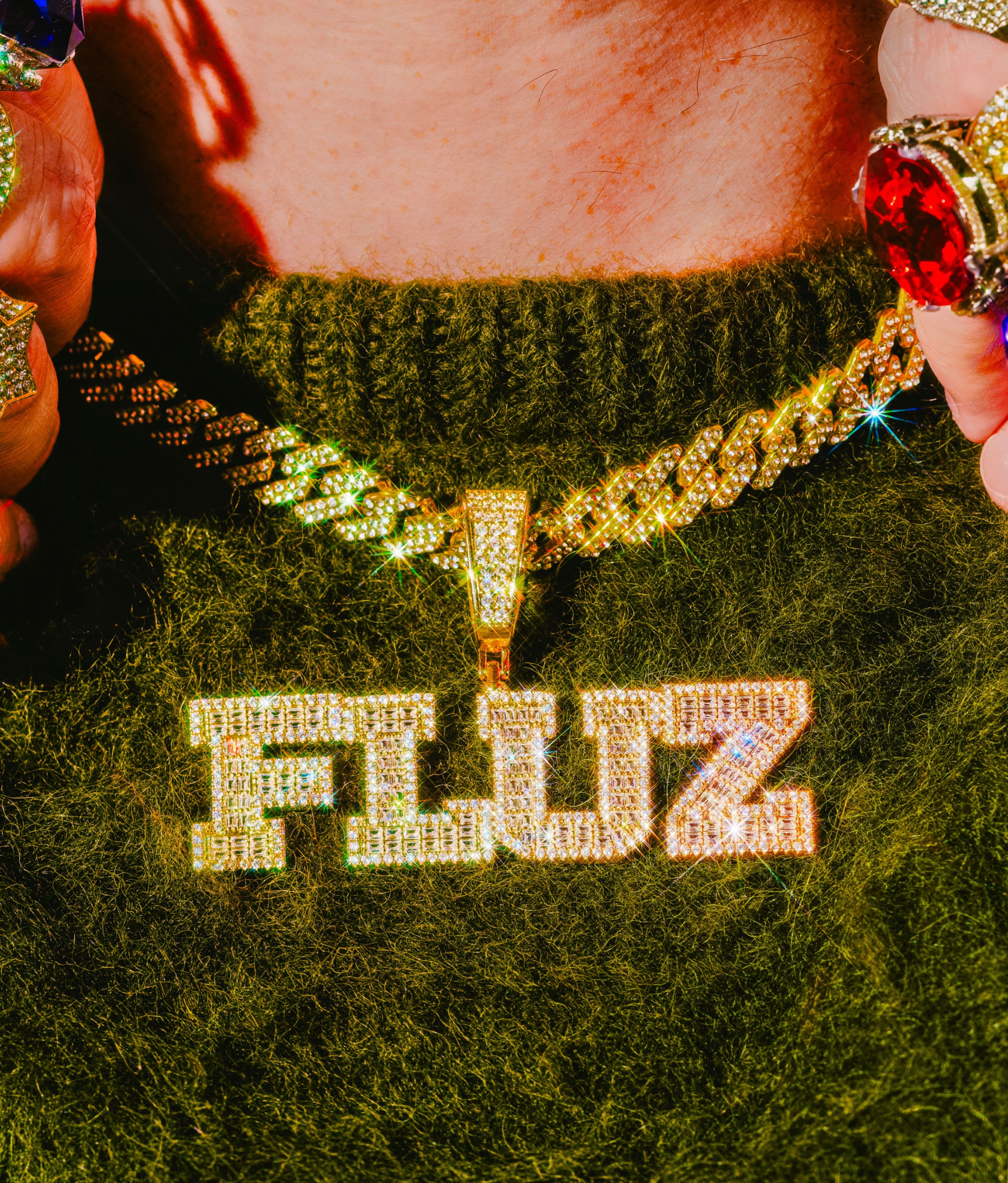 A person wearing a fuzzy green sweater displays a gold chain necklace with a large, sparkling pendant that reads FLUZ. Both hands are adorned with flashy, colorful gemstone rings.