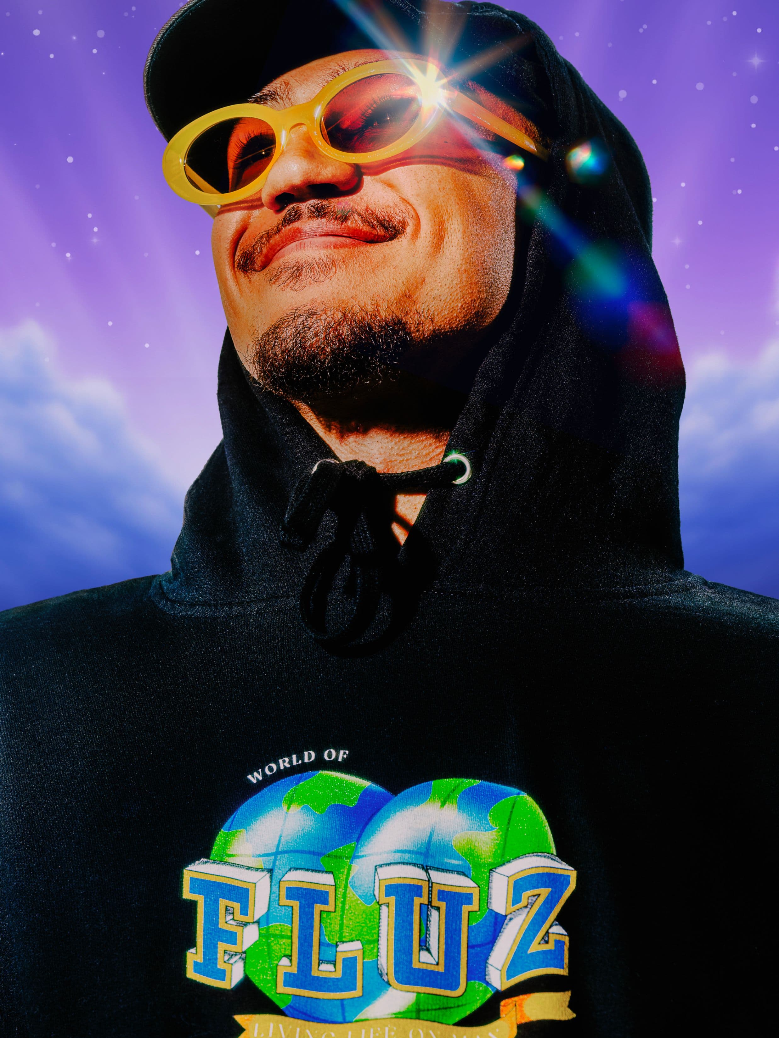 A smiling person in a black hoodie and yellow sunglasses stands against a dreamy, purple sky with stars and clouds. Sunlight glints off the sunglasses, and the hoodie reads World of FLUZ with a globe graphic.