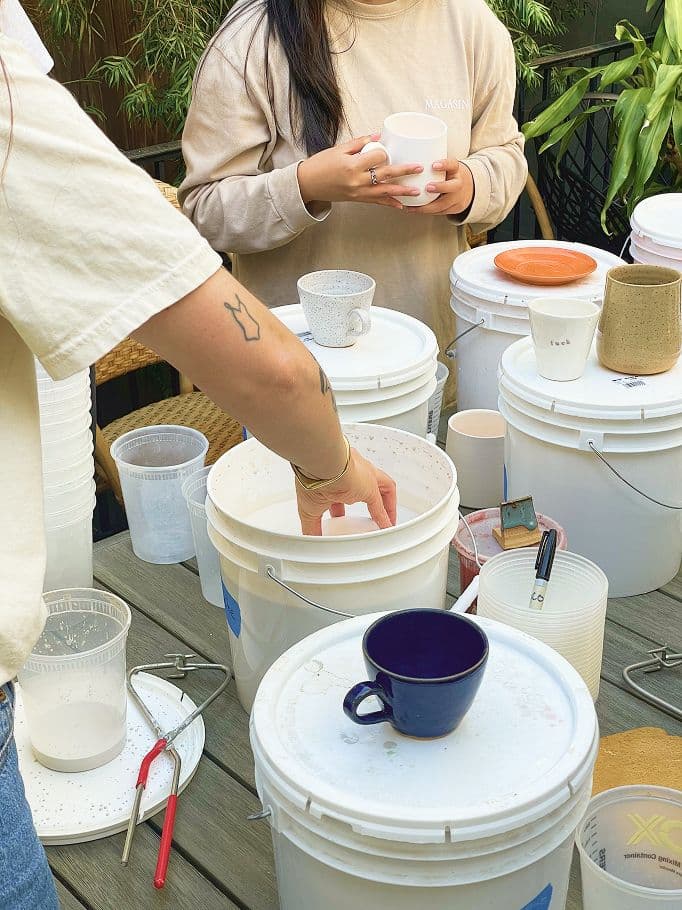 Two people work at a table covered with white buckets, cups, and paint brushes. One person dips a white cup into a bucket, while the other holds a mug. Various mugs and tools are scattered around.