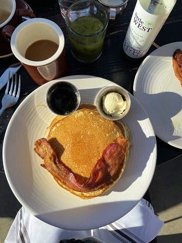A plate with two pancakes topped with a strip of bacon, a small cup of whipped butter, and a small cup of syrup, arranged to resemble a smiling face. Coffee, juice, and bottled drink are nearby.