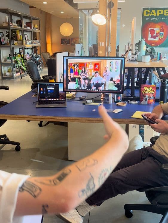 Koto Los Angeles office scene: one person gestures at a laptop and monitor with a group video call, another uses a smartphone; modern office with shelves and posters.