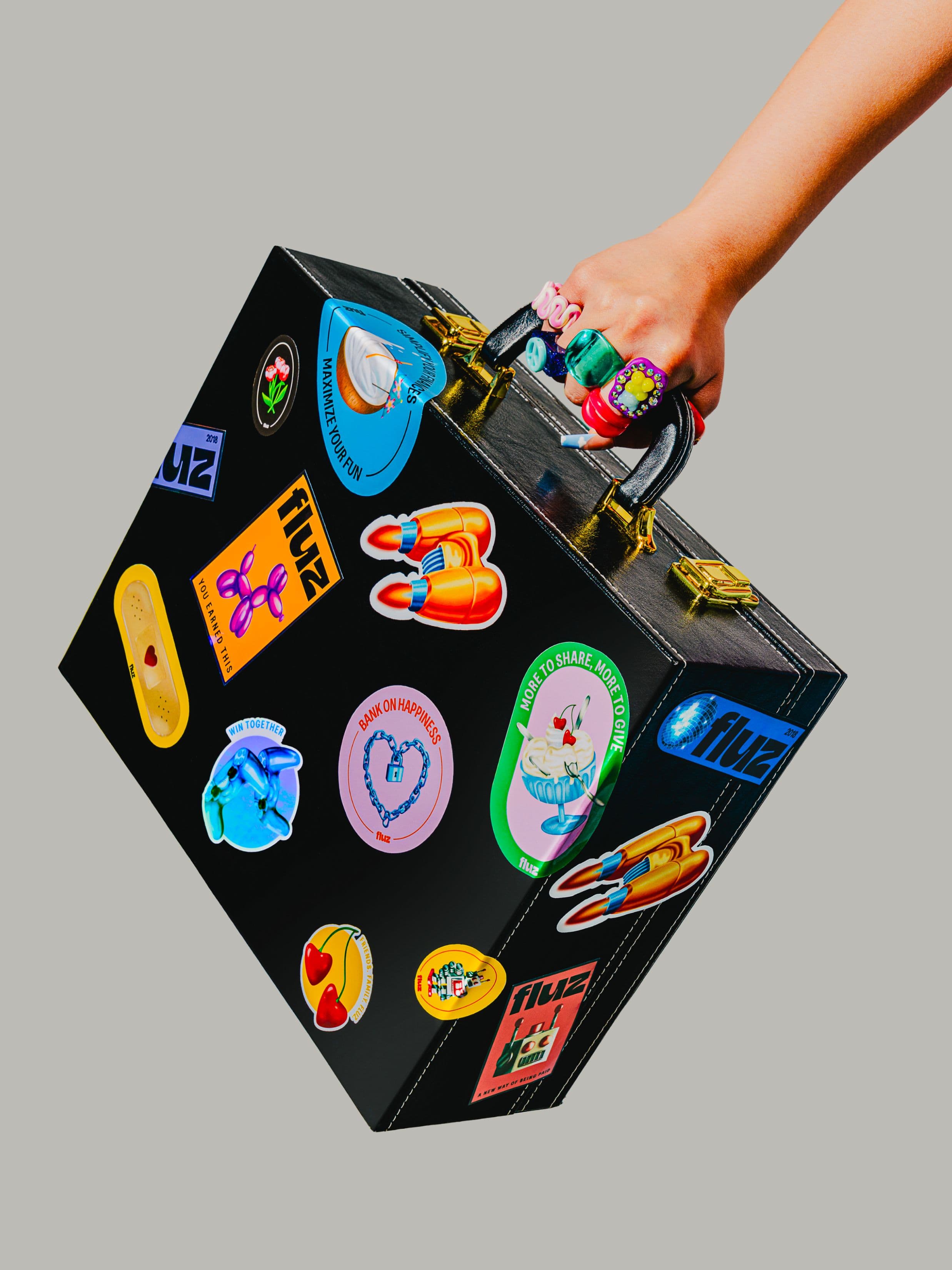 A hand holding a black suitcase decorated with colorful stickers, including hearts, rockets, and various logos.