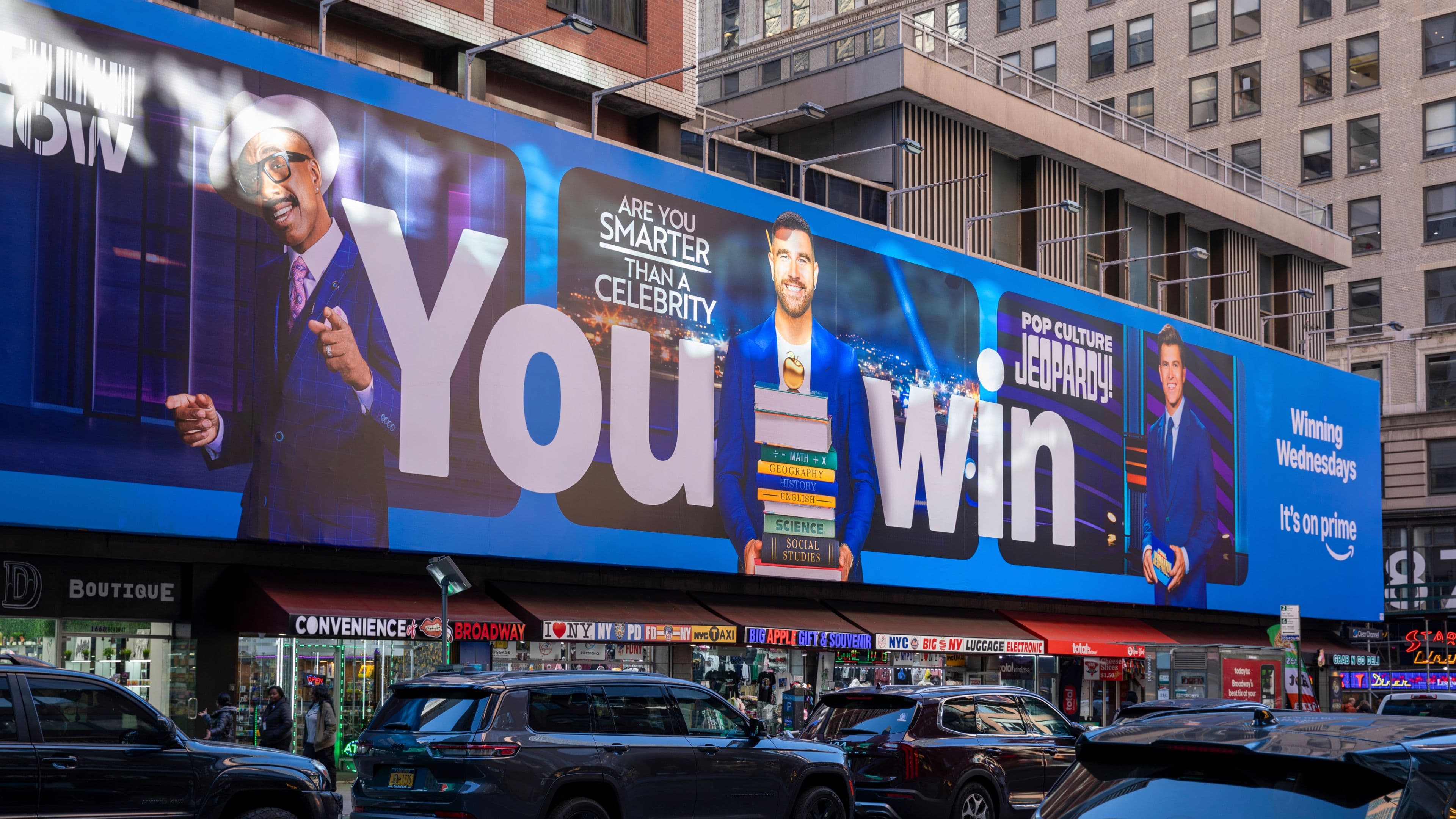 "Pop Culture Jeopardy!" billboard in a city, featuring hosts with trivia cards and books, text about beating celebrities, with street scene below.