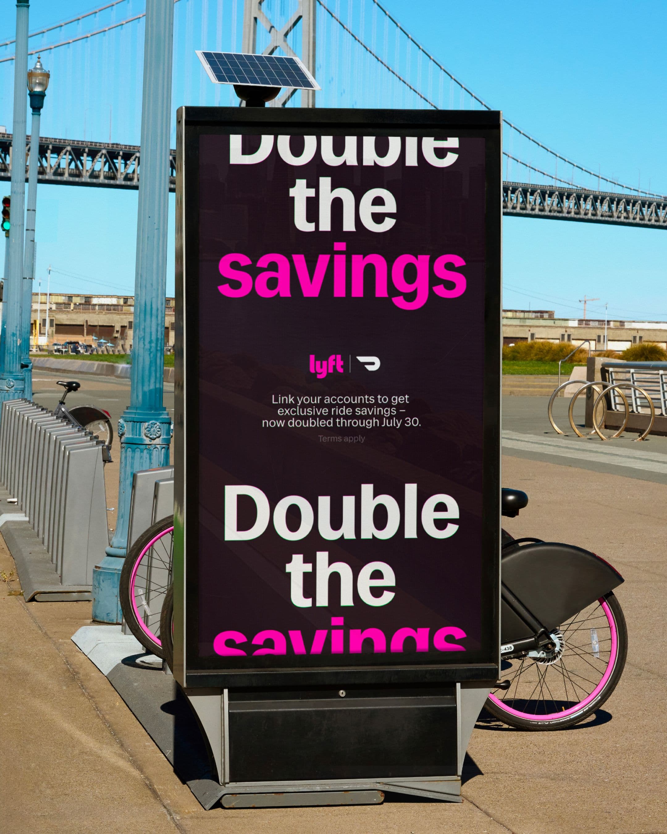 A street advertisement displays the text “Double the savings” in large white and pink letters, promoting a Lyft offer. A pink bike is parked behind the ad, and a bridge is visible in the background.