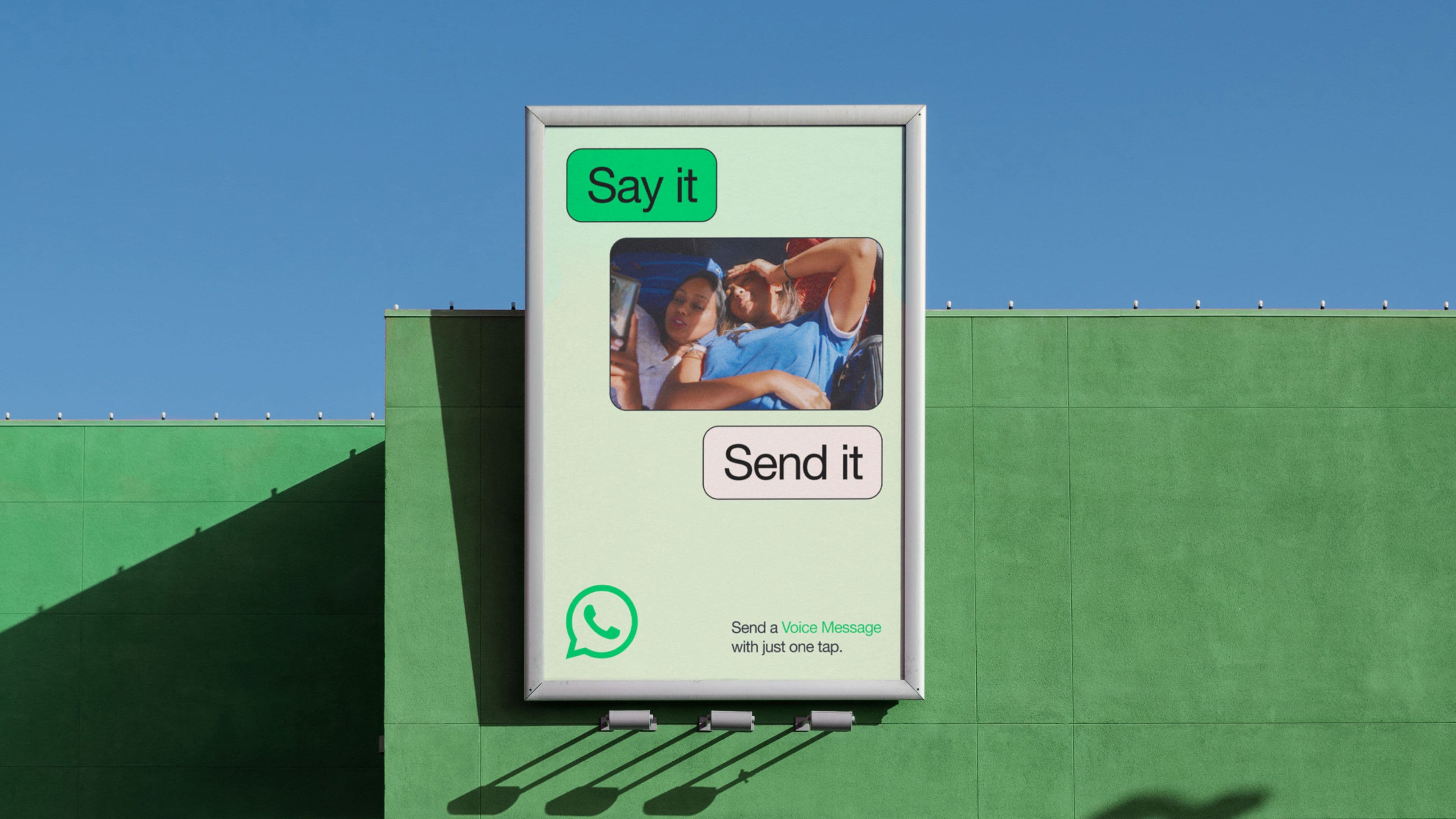 A billboard on a green building shows a WhatsApp ad with the phrases Say it and Send it, along with a photo of two people lying down using a phone. Text encouraging people to sending a voice message appear below.