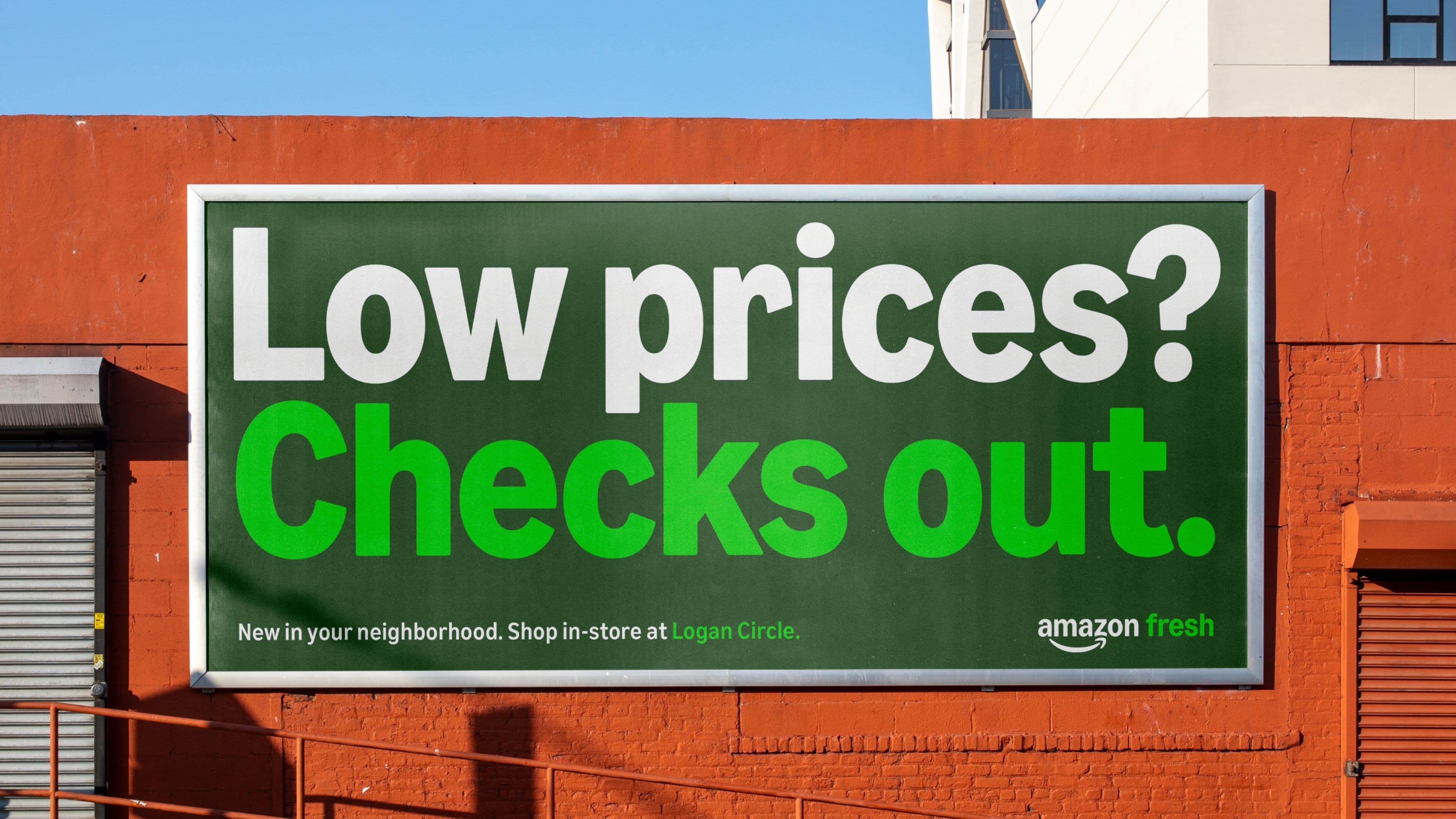 A green Amazon Fresh billboard on a red brick wall reads, "Low prices? Checks out". Smaller text below says, New in your neighborhood.