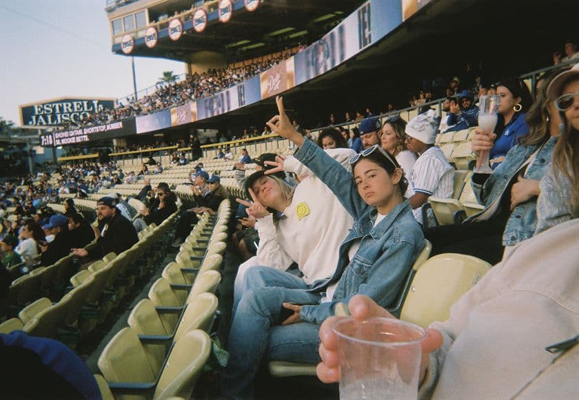 Two people sit in stadium seats among a crowd, both making peace signs and smiling at the camera.