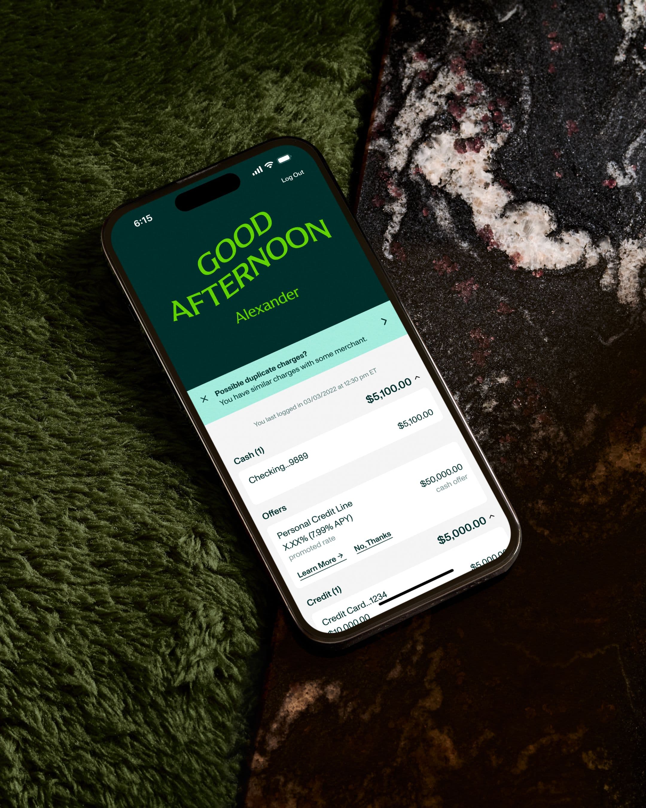Huntington Bank app on a smartphone showing greeting and account balances, resting on a green rug by a dark marbled surface.