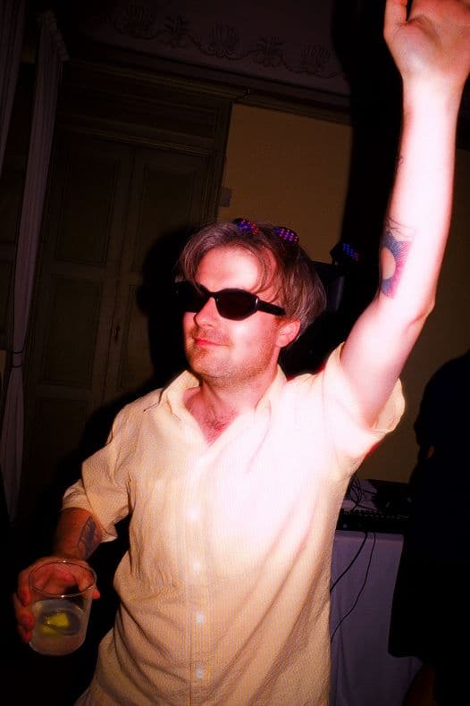 Man wearing sunglasses and yellow shirt at a party, holding a drink with one arm raised, colorful lights around him.